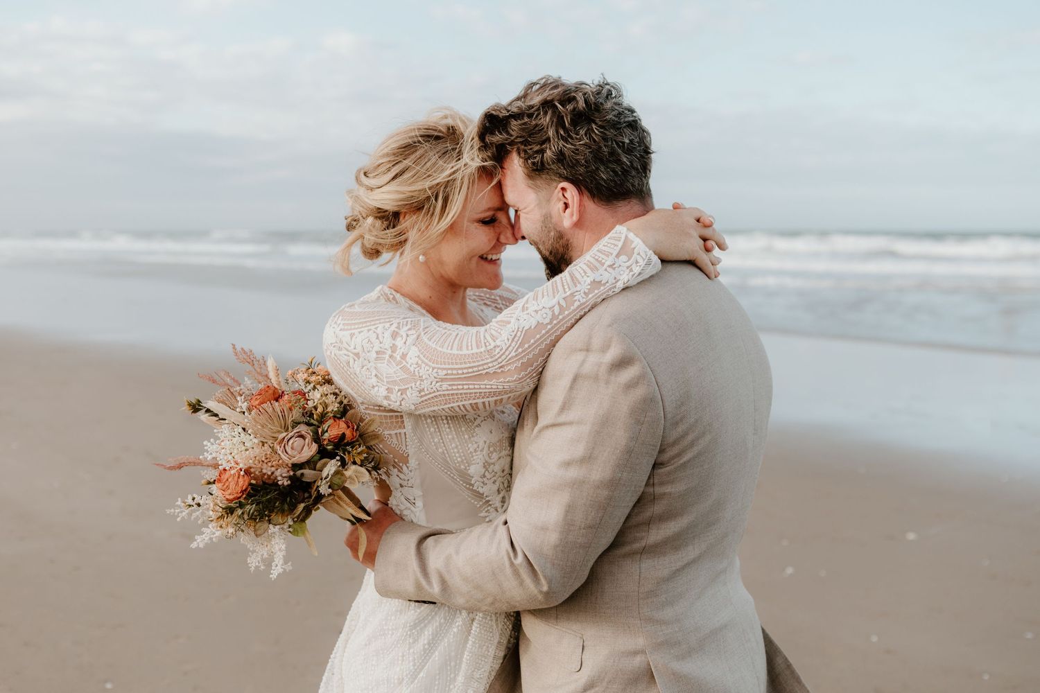 A newlywed couple share a moment embracing on the sand as waves roll in behind them. Tauranga wedding photographer