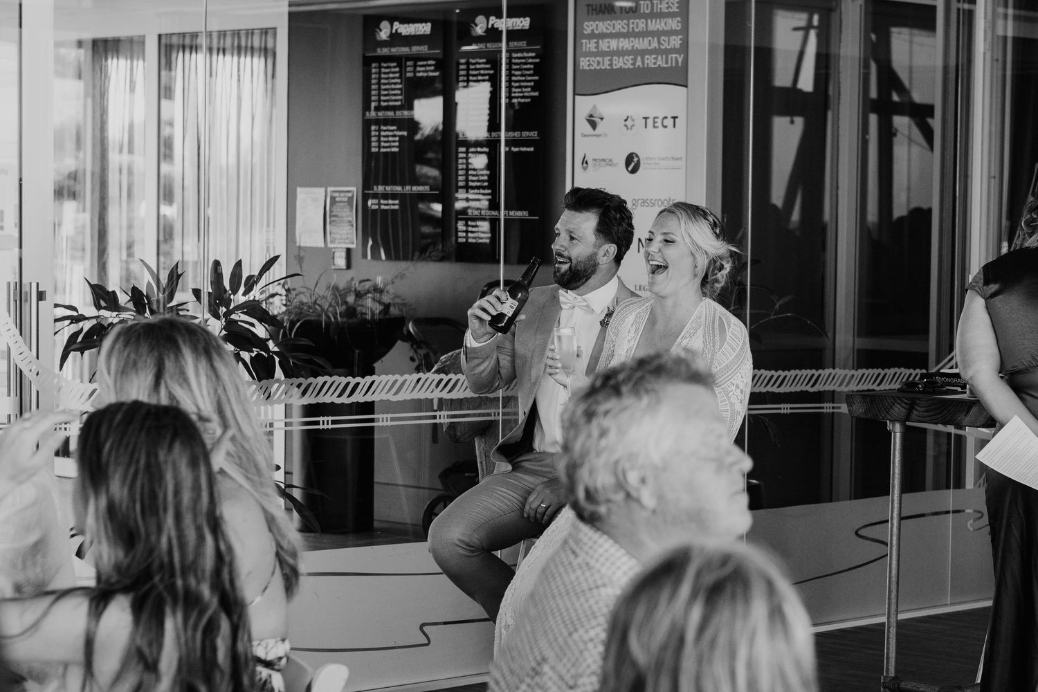 Candid black and white photo of wedding guests socializing at an indoor venue. Papamoa Surf Lifesaving Club Wedding