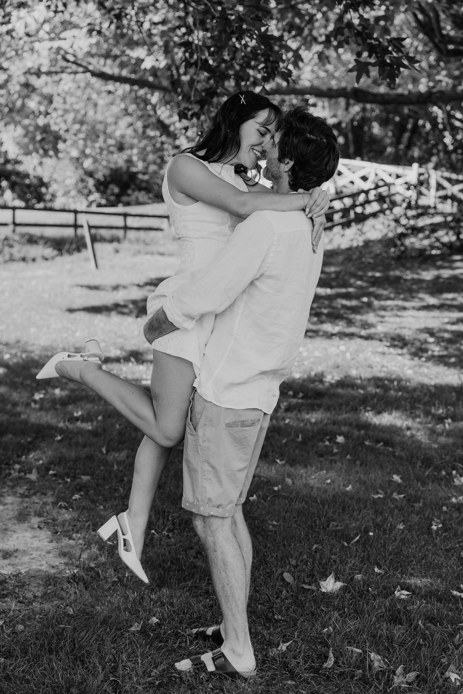 Black and white photo of a couple embracing and kissing outdoors, man lifting woman in a park setting.