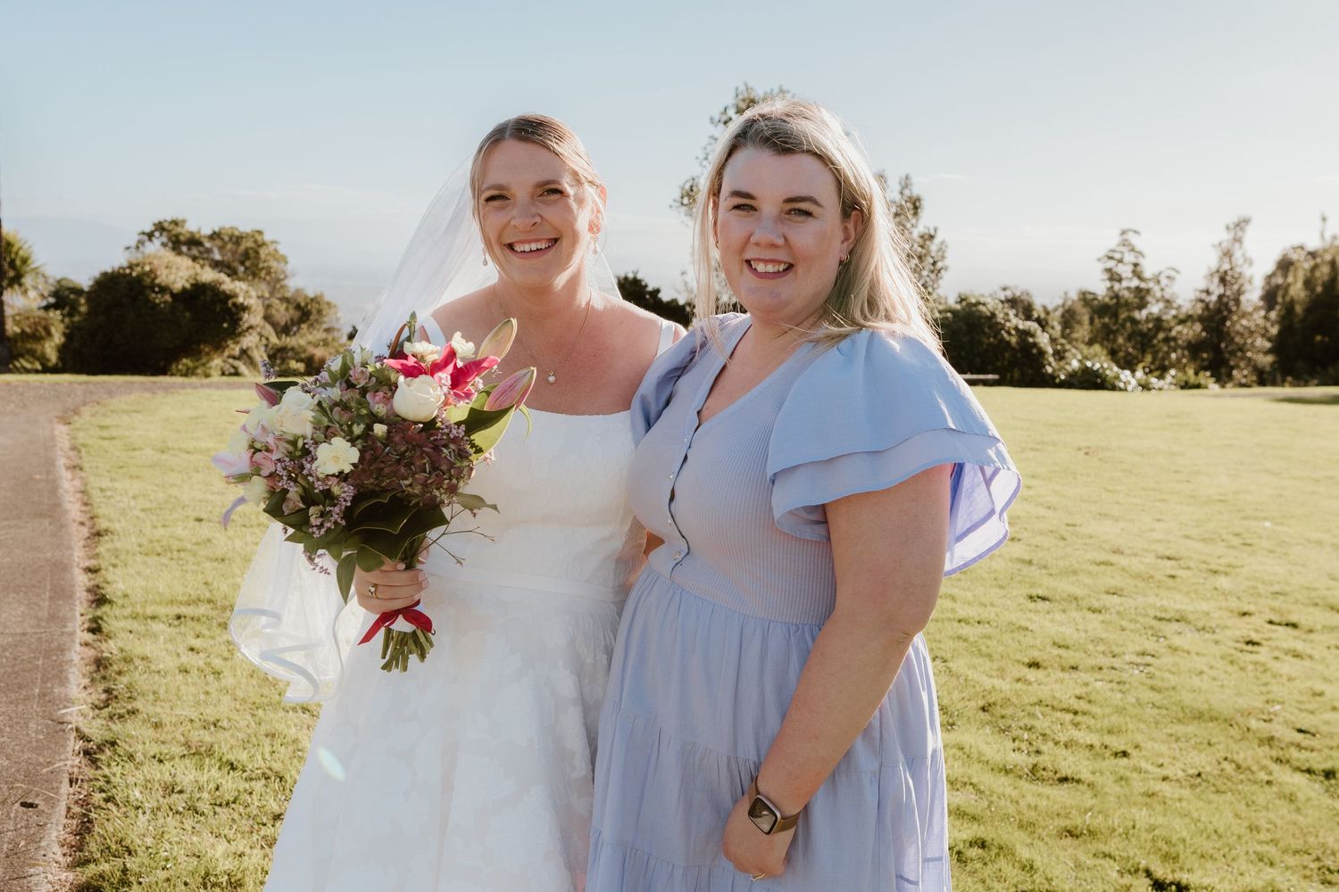Wedding party members in light blue dresses pose together in a sunlit outdoor field with bouquets.