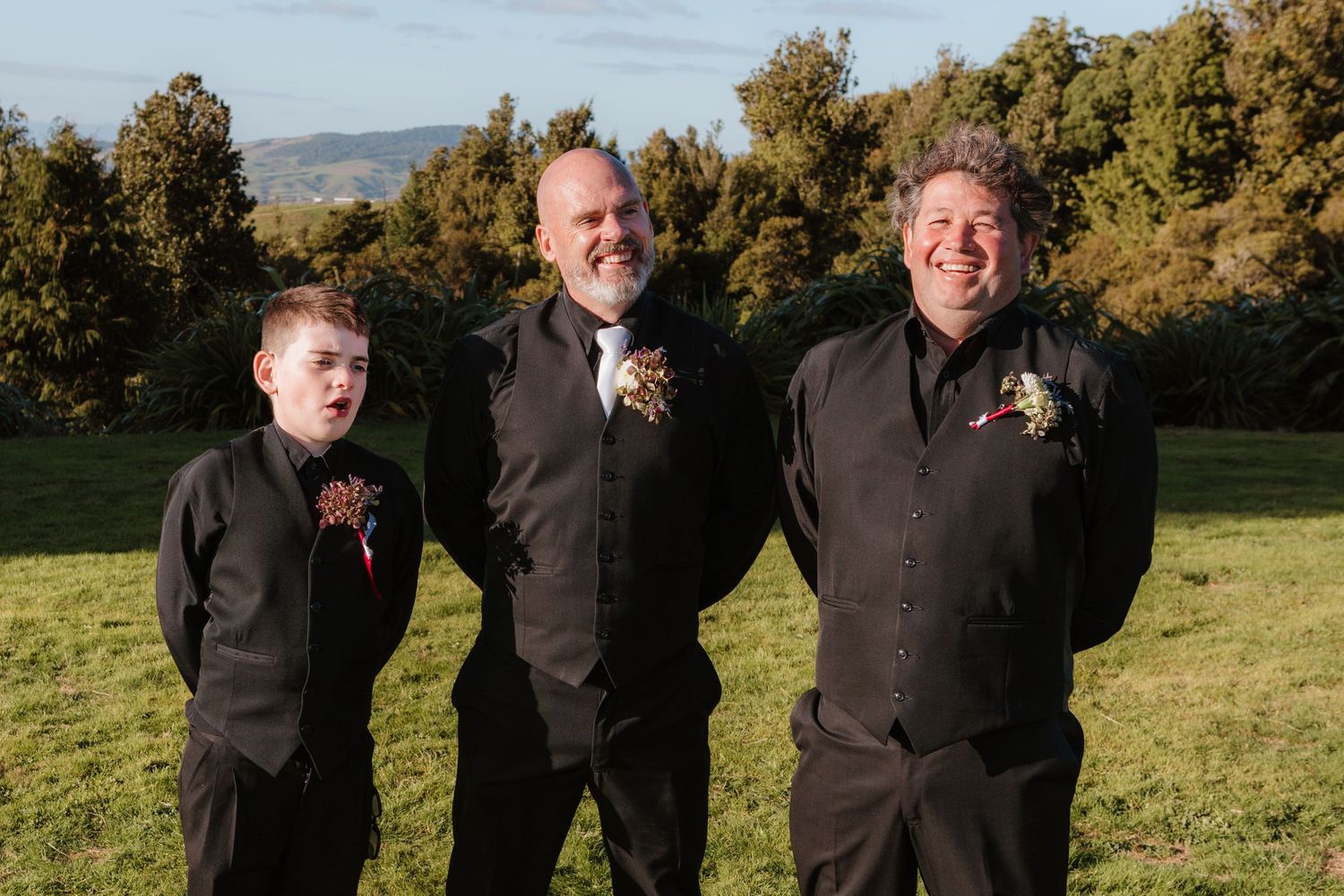Three people in matching black formal attire stand together smiling on a grassy hillside with trees in background.