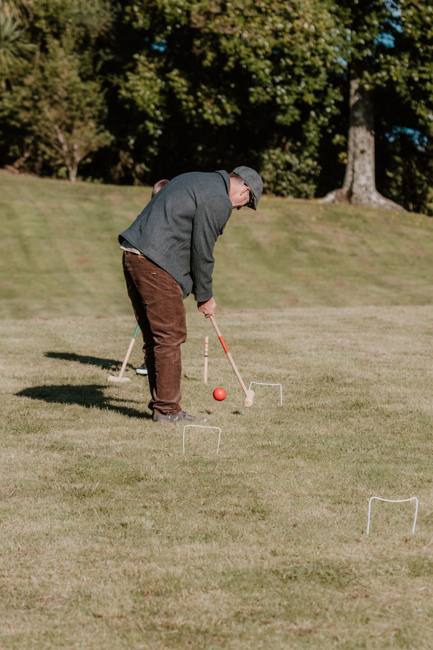 A person in a gray sweater and brown pants plays croquet on a sunny grassy lawn with hoops set up on the field.