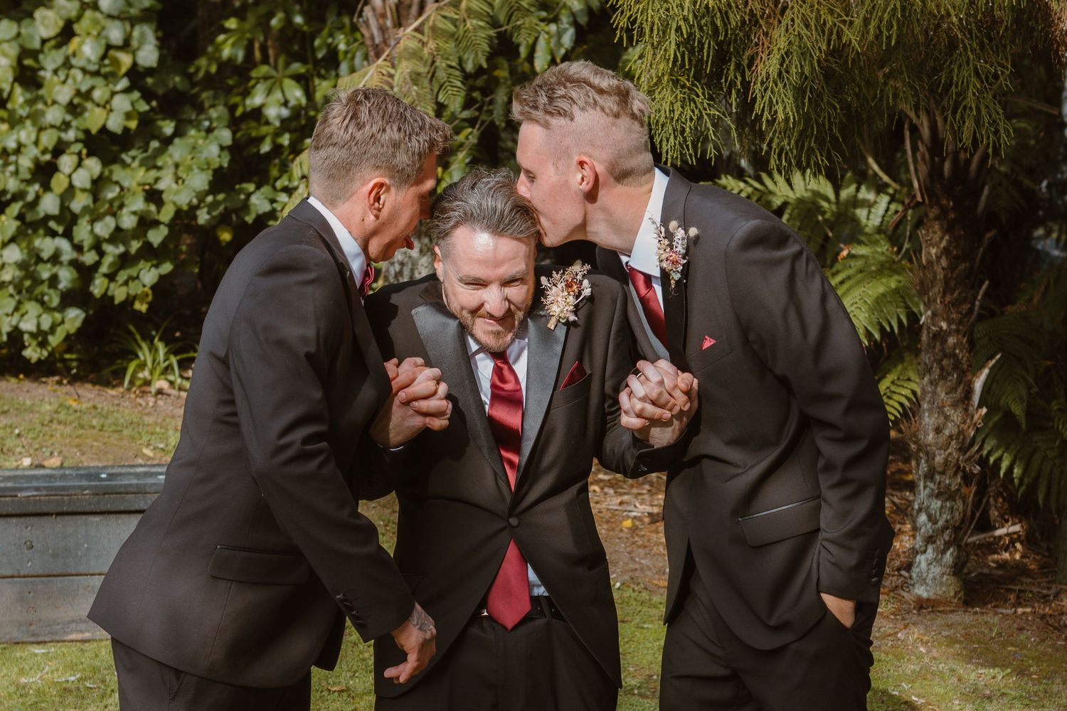 Three people in dark suits share a lighthearted moment at an outdoor event with lush greenery in the background.