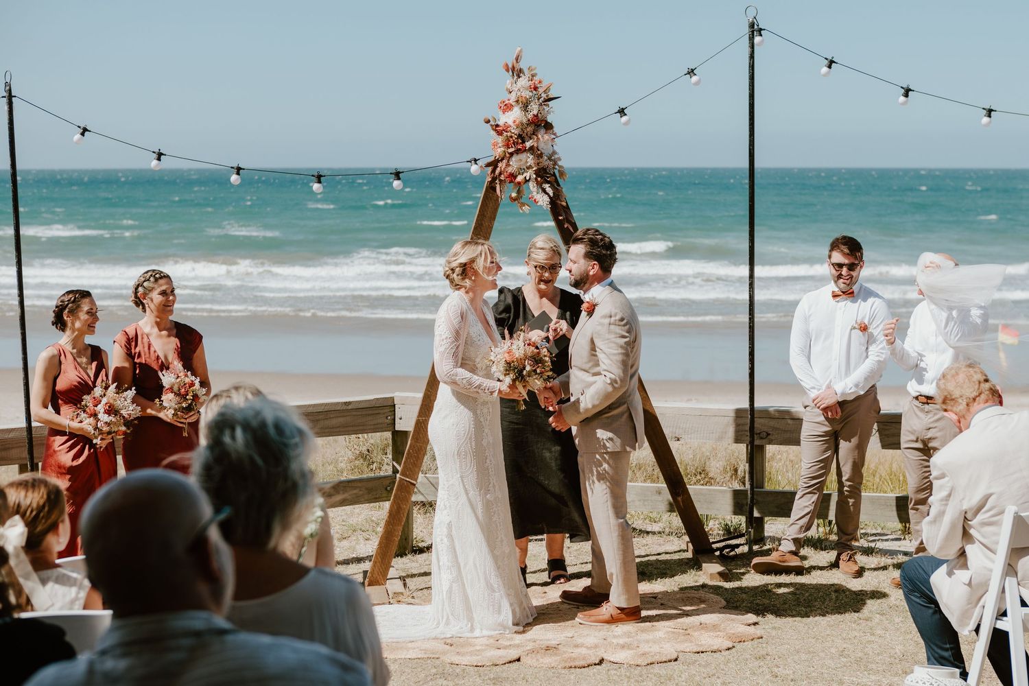 A beachside wedding ceremony takes place under a rustic arch with string lights and waves crashing in the background.