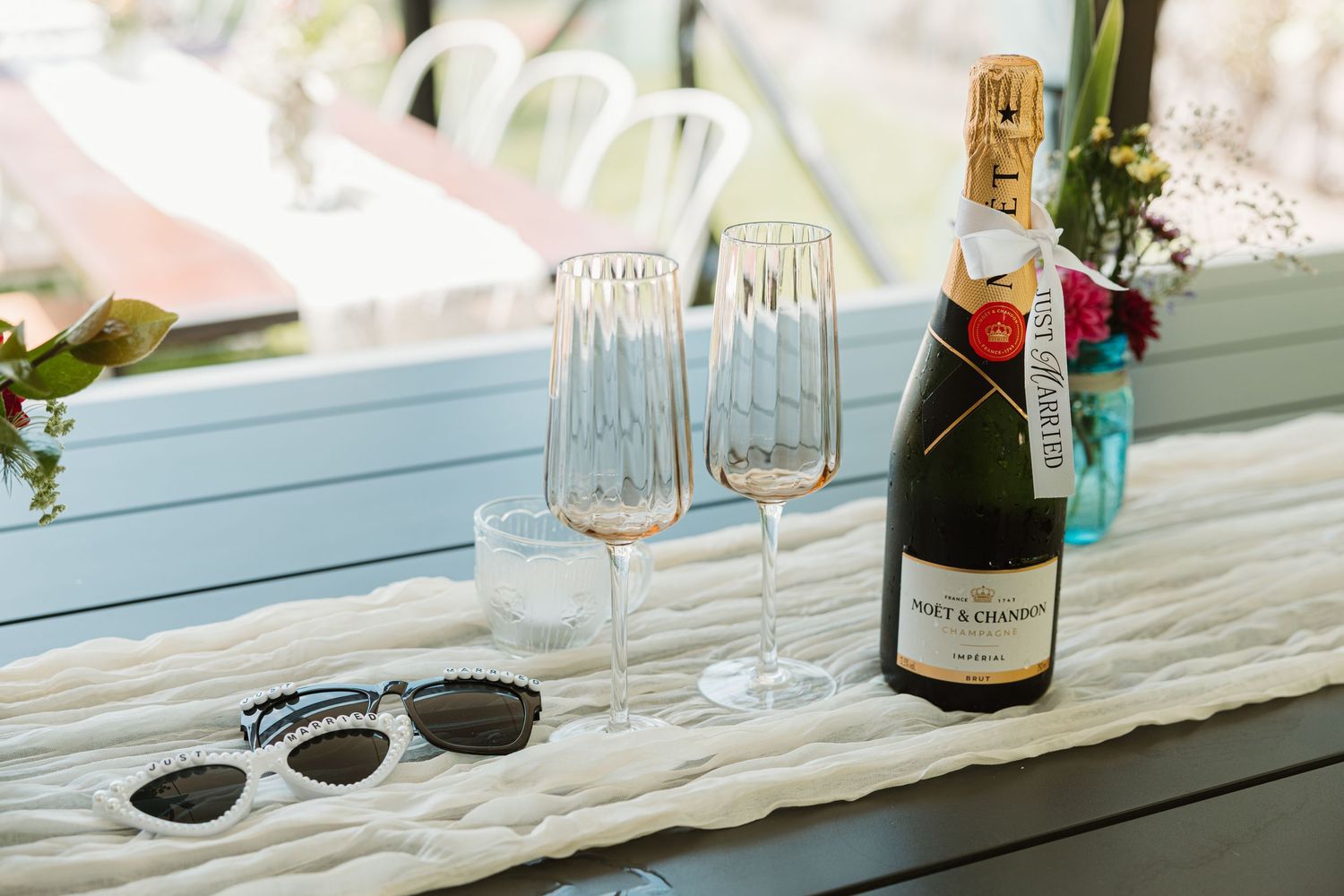 Moët & Chandon champagne bottle with two crystal flute glasses and sunglasses on a white blanket near a window.