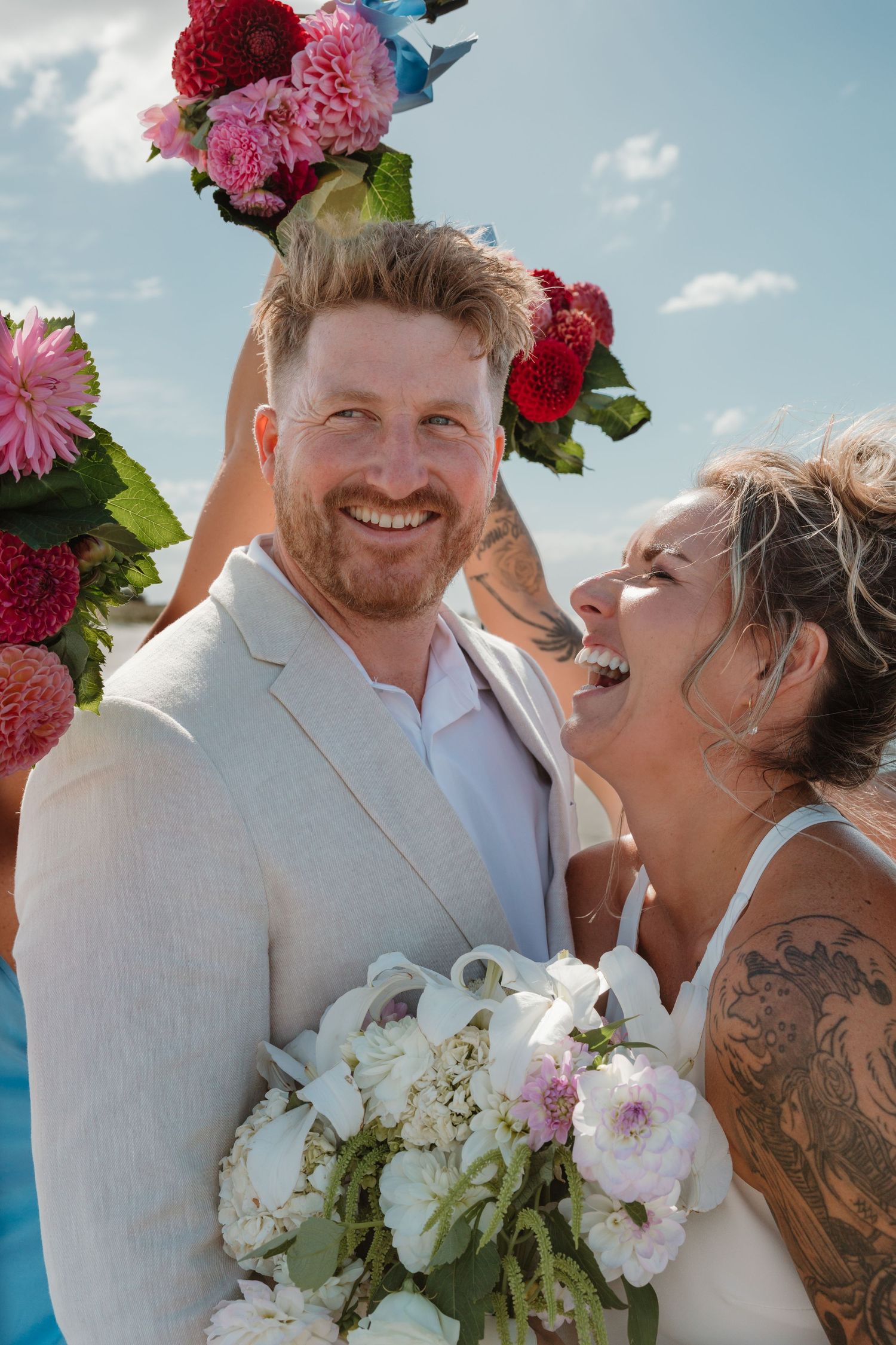 Joyful bride with tattoos holding white bouquet and groom in tan suit laughing under colorful floral arch outdoors.