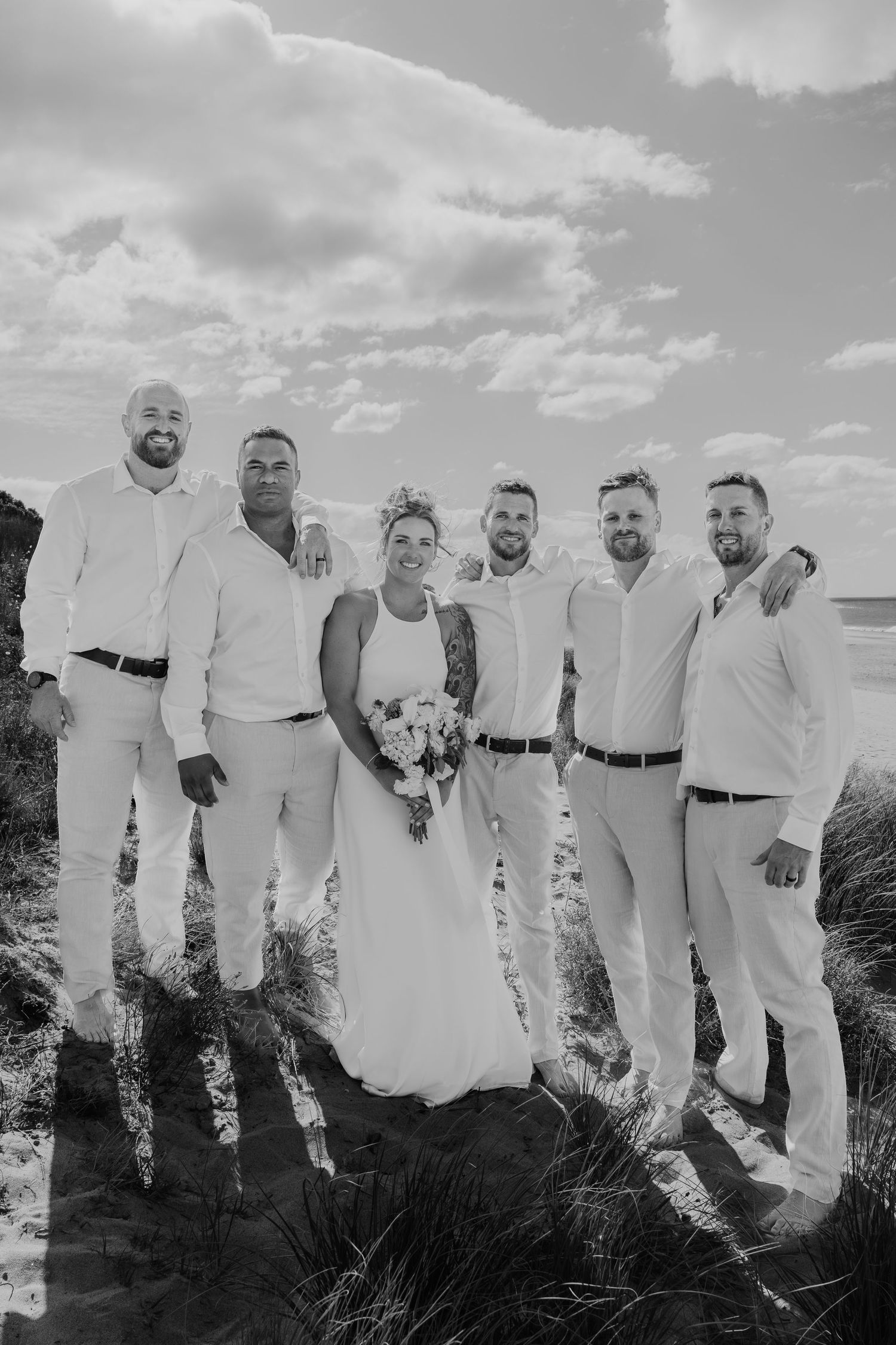 Black and white photo of a bride holding a bouquet posing with five groomsmen on a sandy beach.