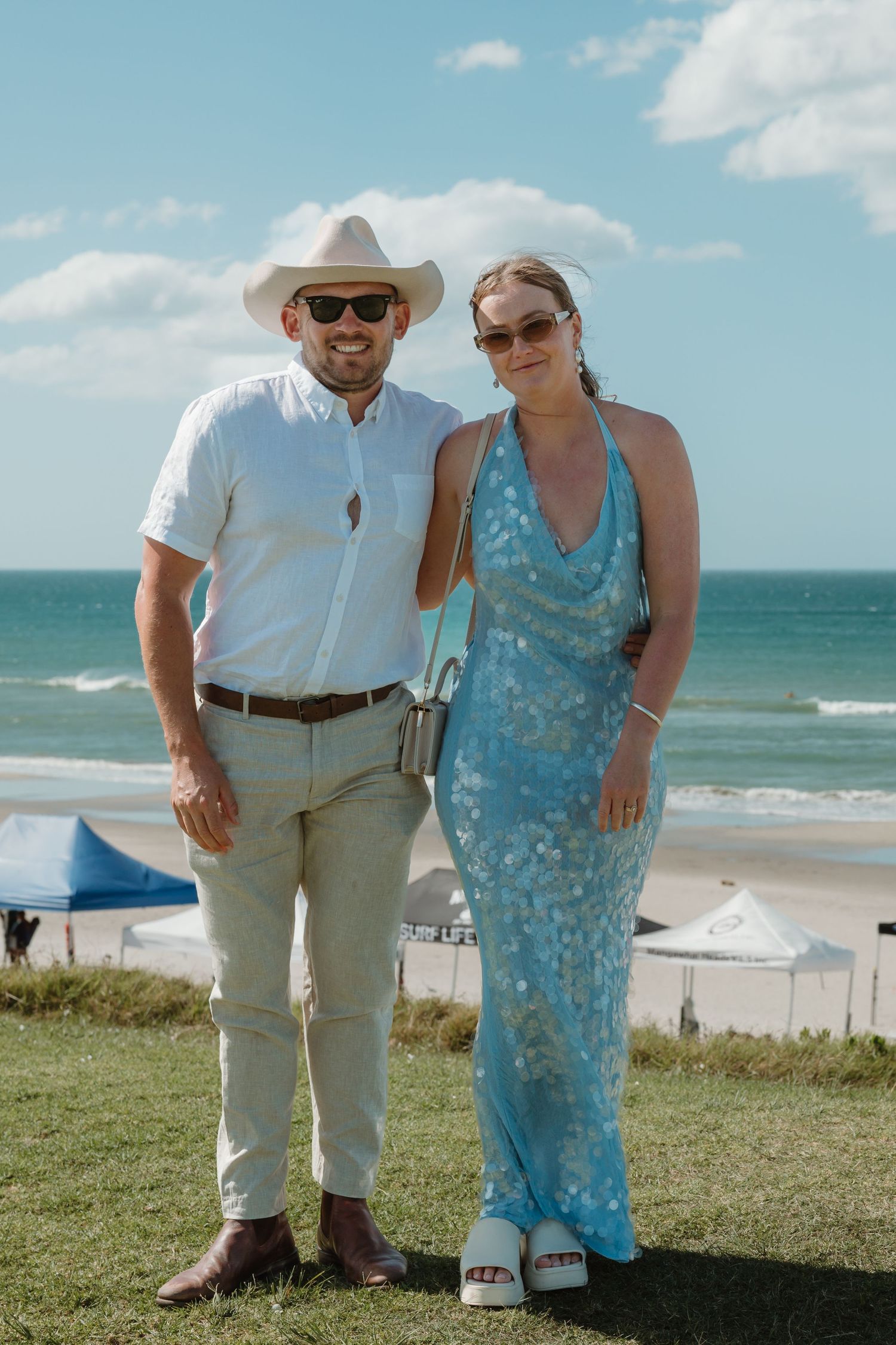 Couple posing together on a sunny beachside lawn, man in white shirt and cowboy hat, woman in blue floral maxi dress.