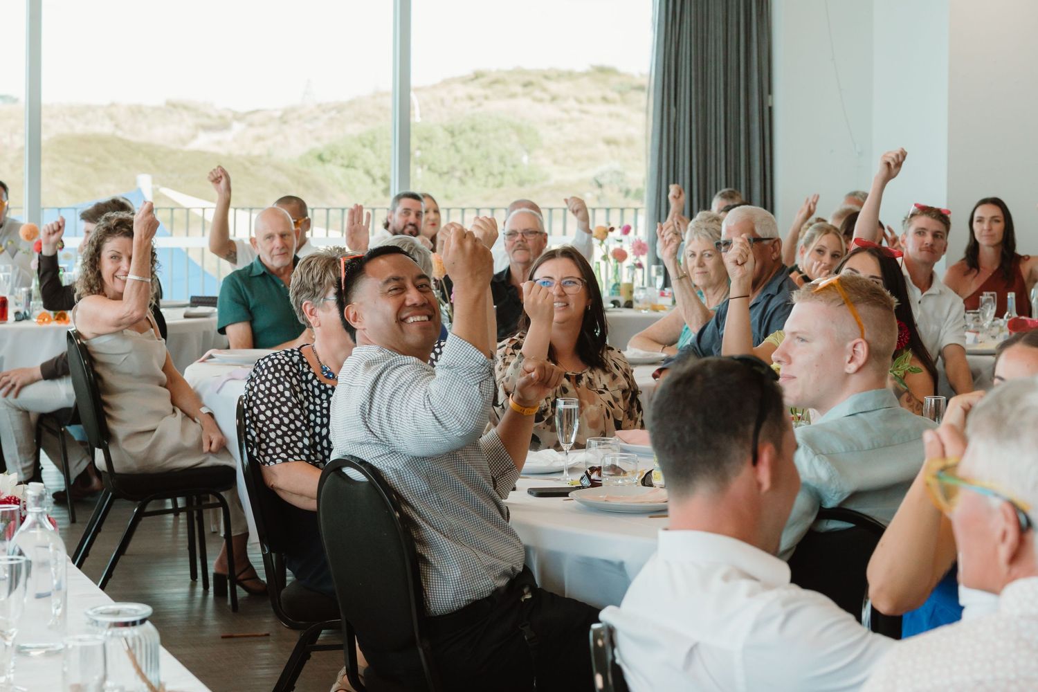 Guests cheering and celebrating at a bright seaside restaurant during a lively event or reception.
