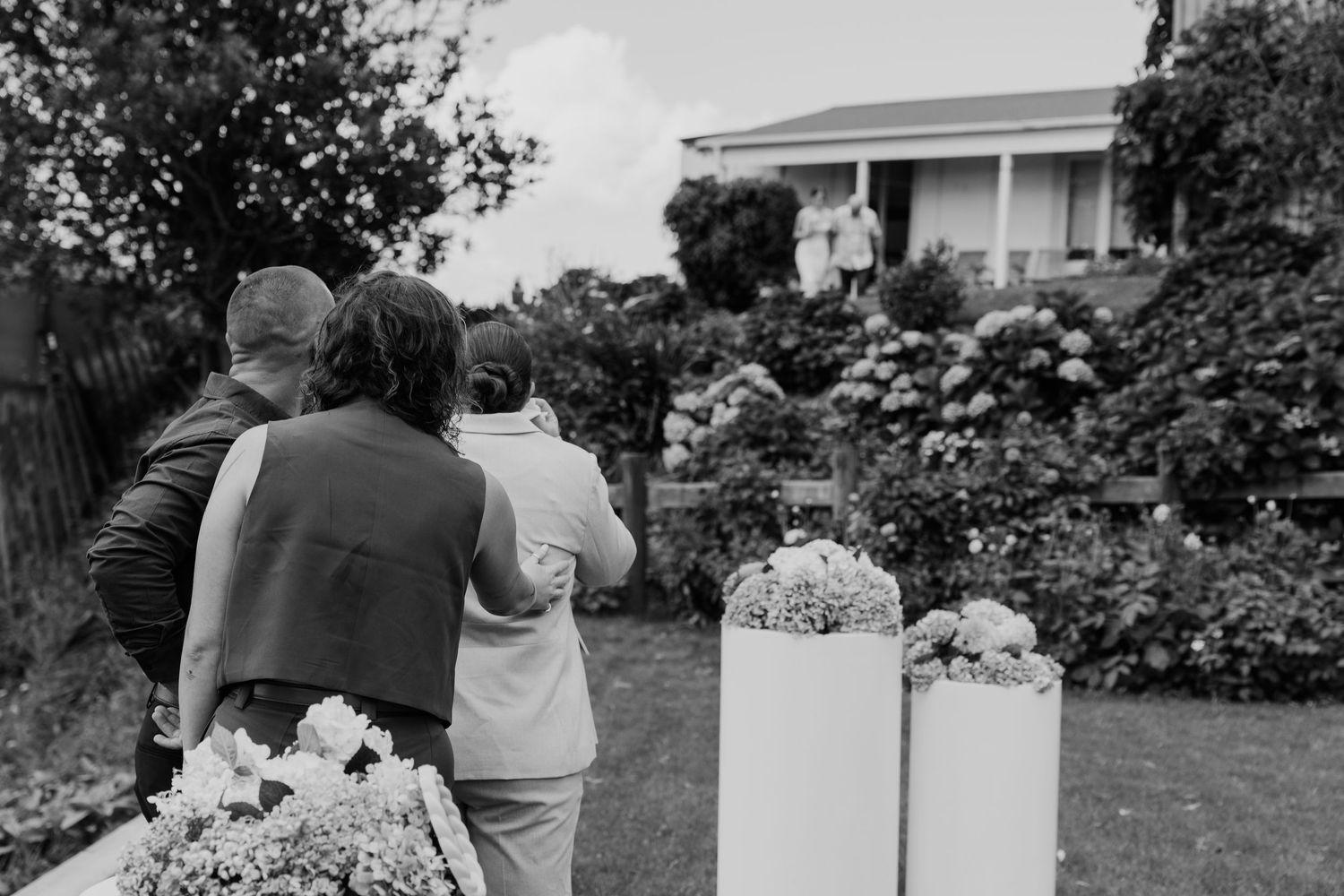 Black and white photo of guests embracing at an outdoor garden wedding ceremony with floral arrangements.