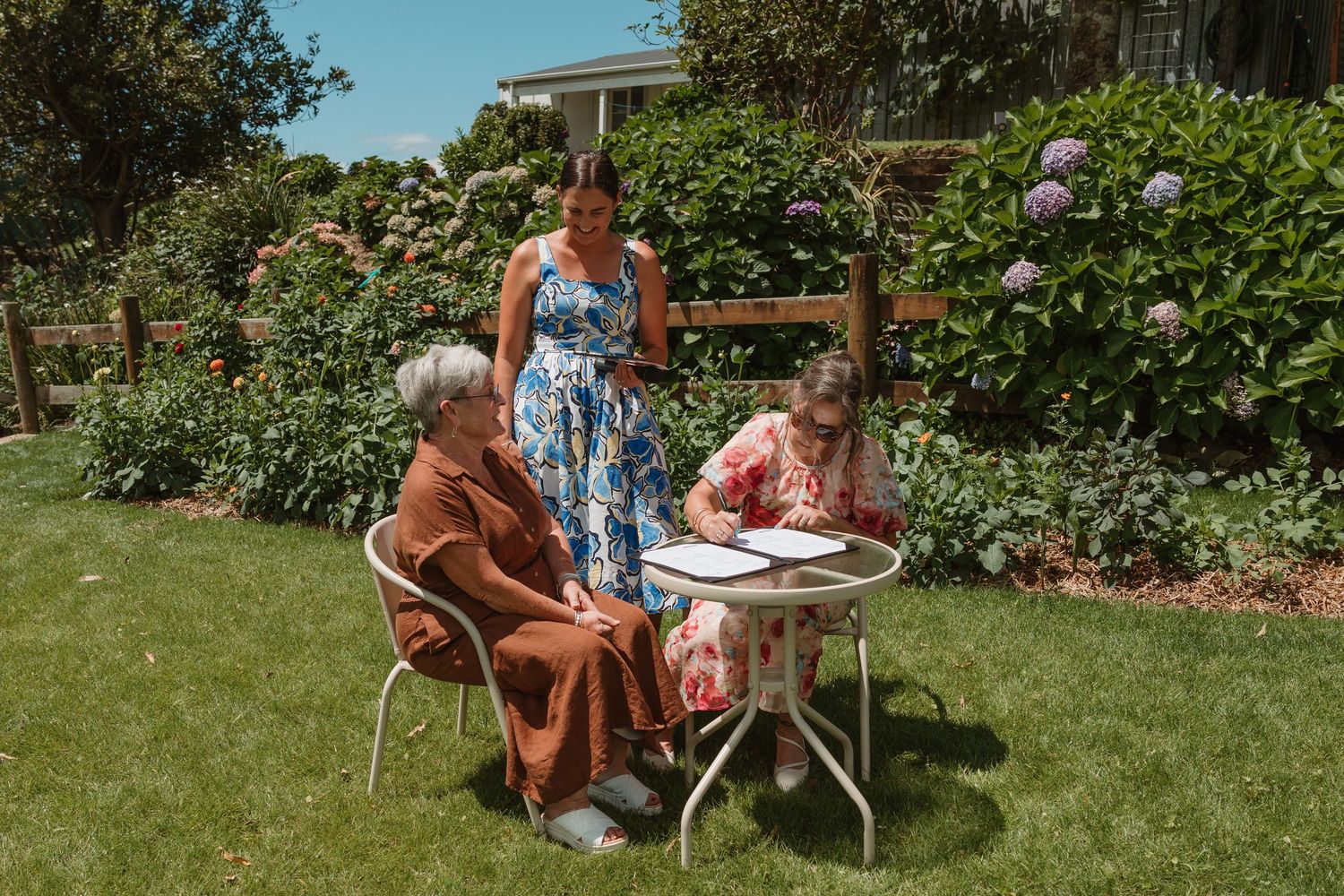 Three women gather around a white garden table on a sunny day, surrounded by lush green hedges and colorful flowers.