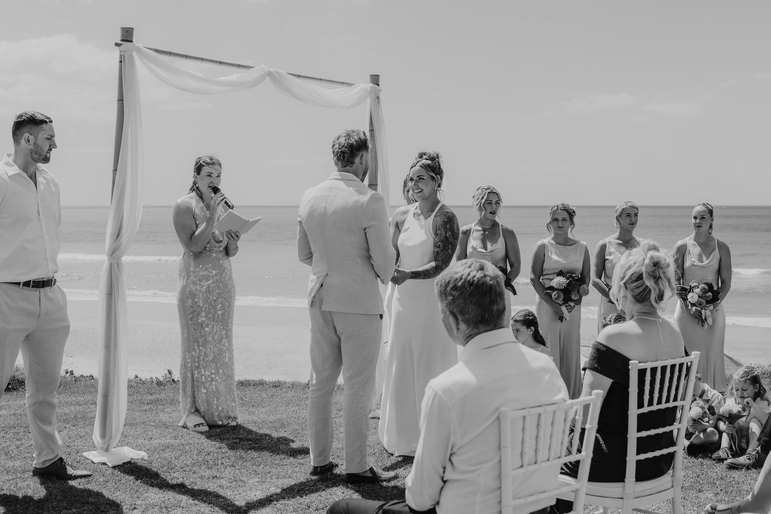 Outdoor beach wedding ceremony with bride and groom exchanging vows under a white arch, bridesmaids standing nearby.