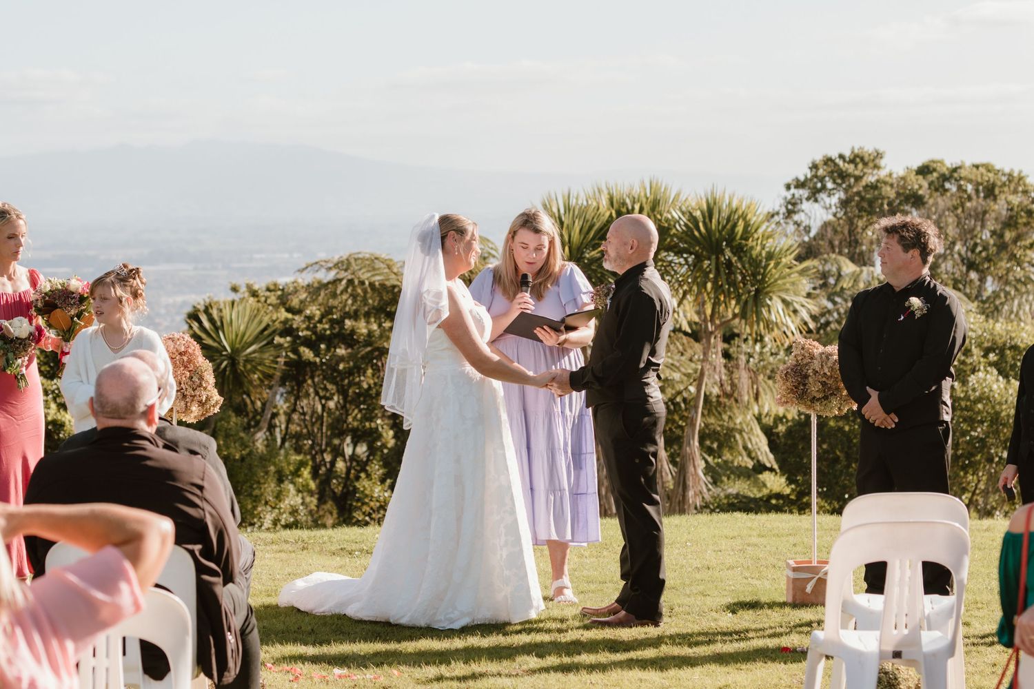 An outdoor wedding ceremony taking place on a hilltop with palm trees and mountains in the background on a sunny day.