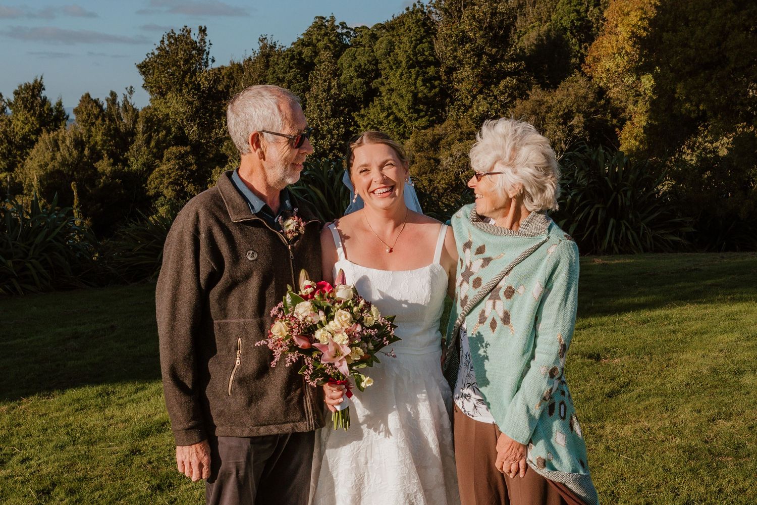 Three people stand together outdoors on a sunny day, with autumn trees in background and a bouquet of flowers.