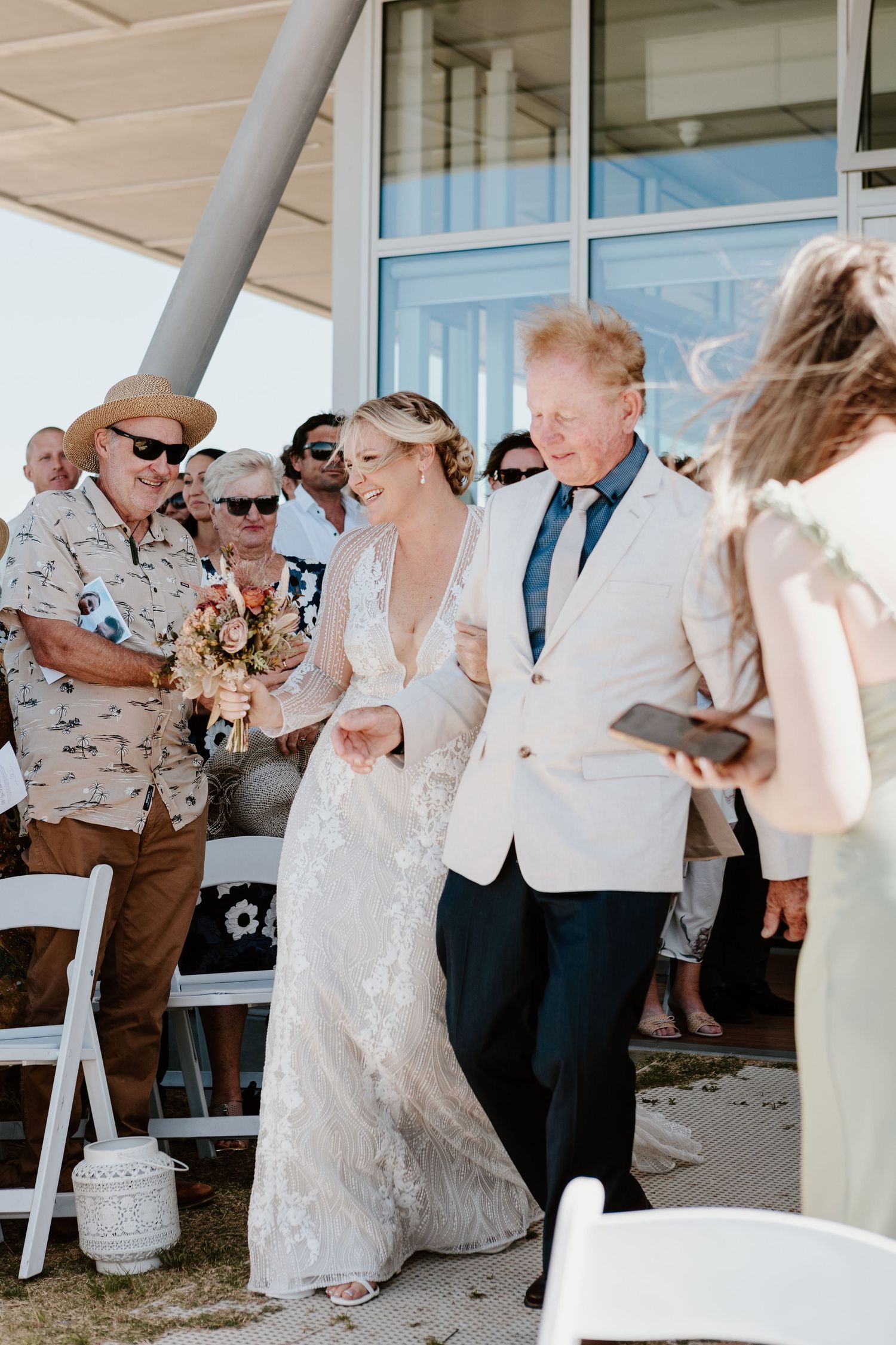 Two people celebrate at an outdoor wedding ceremony as guests look on with white chairs and glass windows in the background.