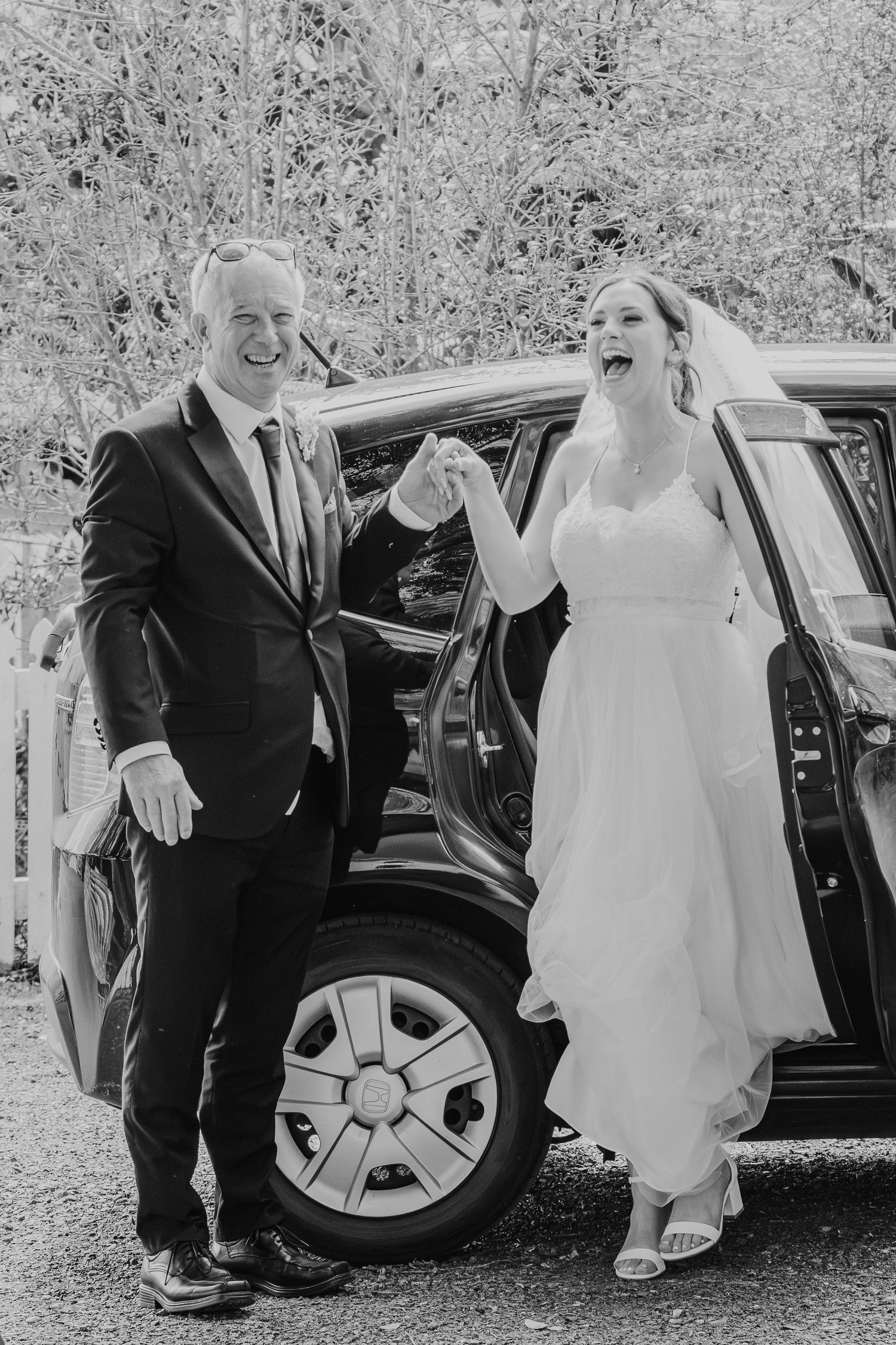 A joyful black and white wedding photo showing a couple laughing together next to a car on their special day.