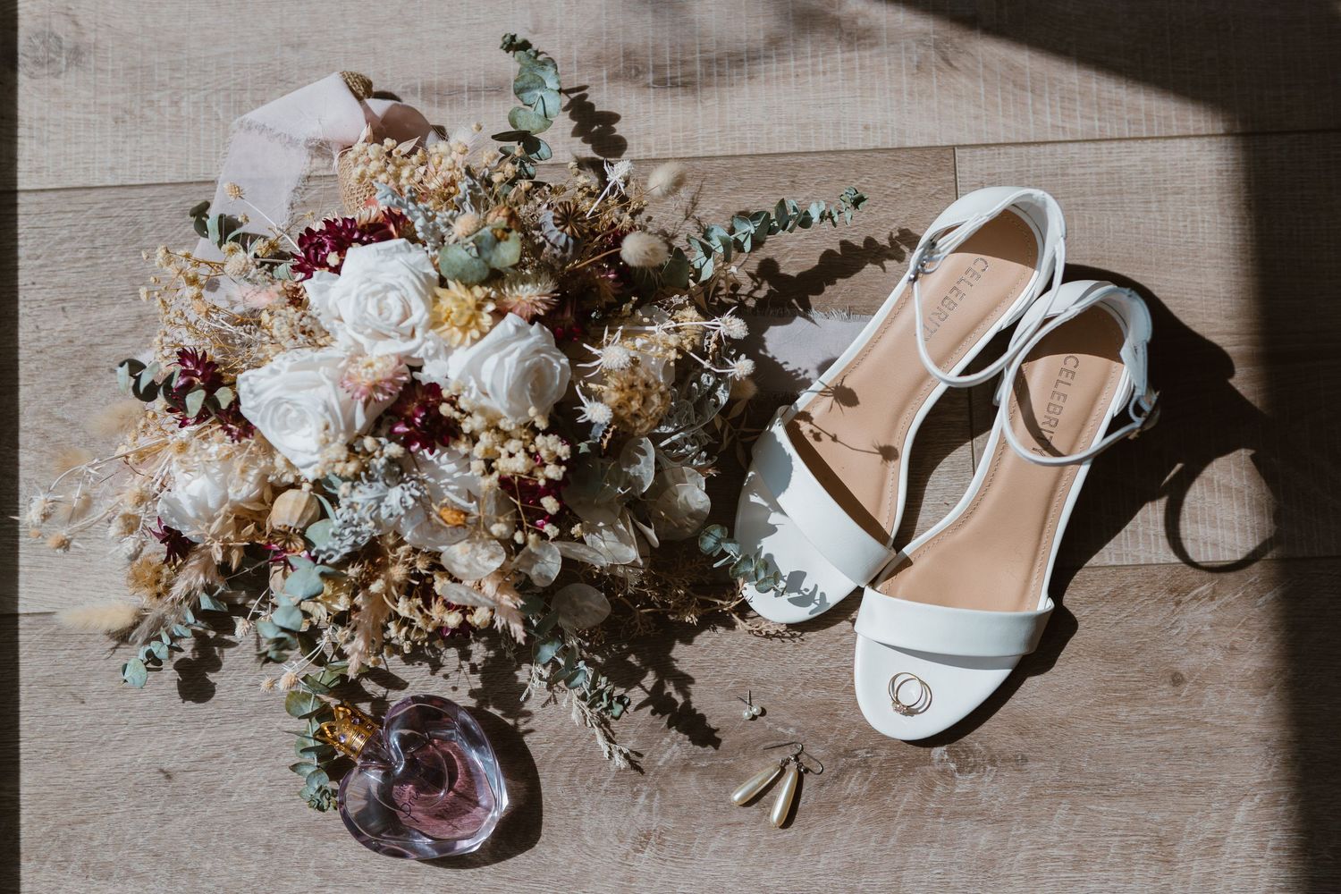Elegant white bridal shoes arranged with dried floral arrangements and delicate tulle fabric on wooden surface.