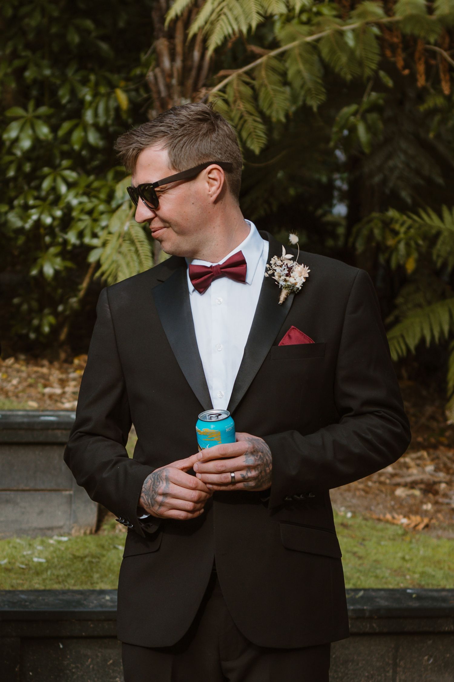 A groom in a black tuxedo with red bow tie and sunglasses holds a turquoise drink can during an outdoor wedding.