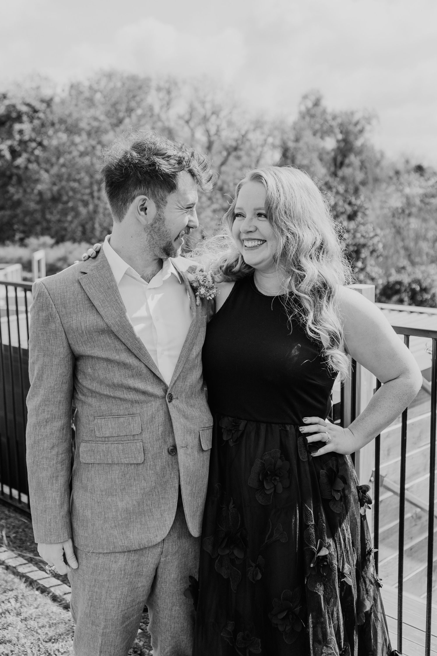 A black and white photo of a couple laughing together outdoors, her in a black dress and him in a gray suit.
