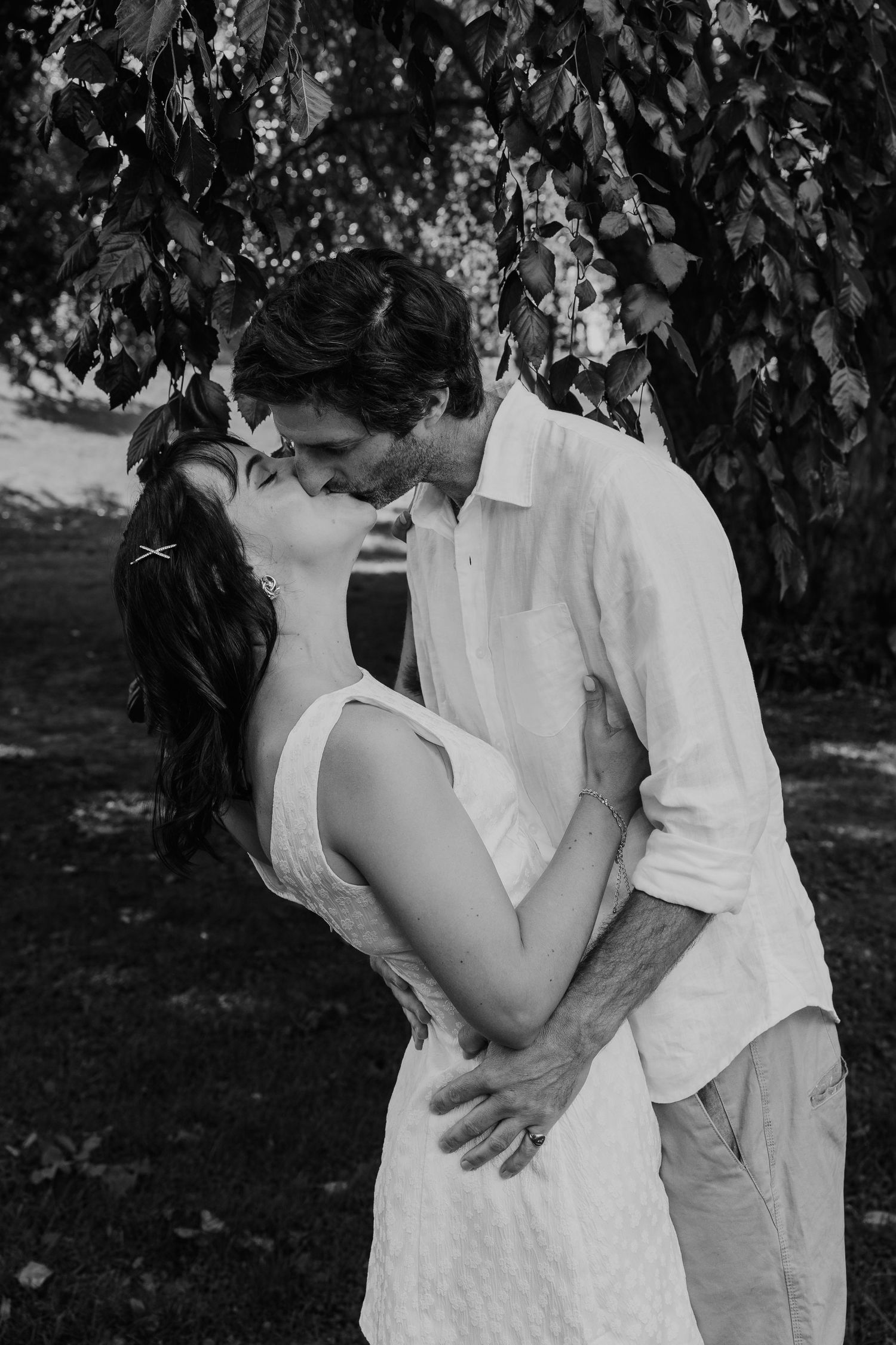 Black and white photo of a couple kissing under leafy trees, dressed in white. Elopement photographer in Tauranga, Bay of Plenty