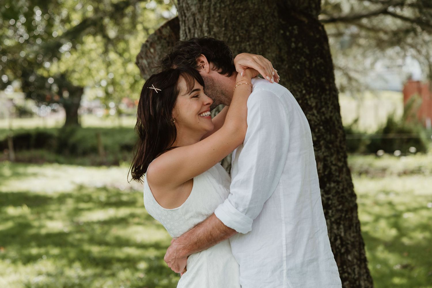 Couple embracing romantically by a tree in a lush green park, both dressed in white outfits.