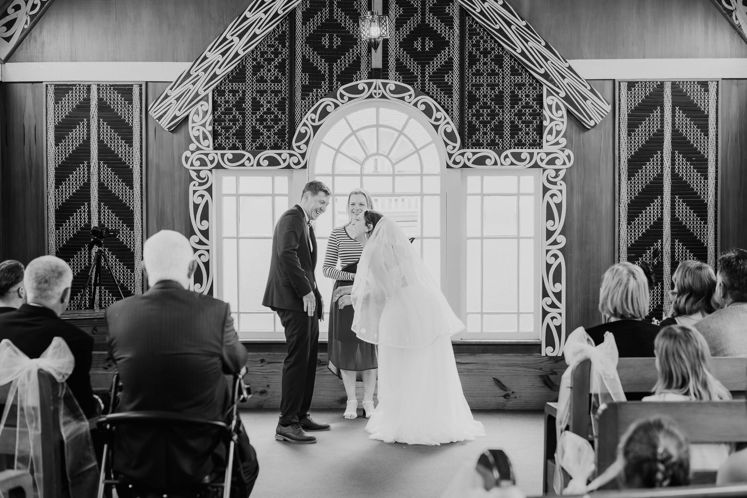 Black and white wedding ceremony photo with guests seated beneath ornate architectural details.