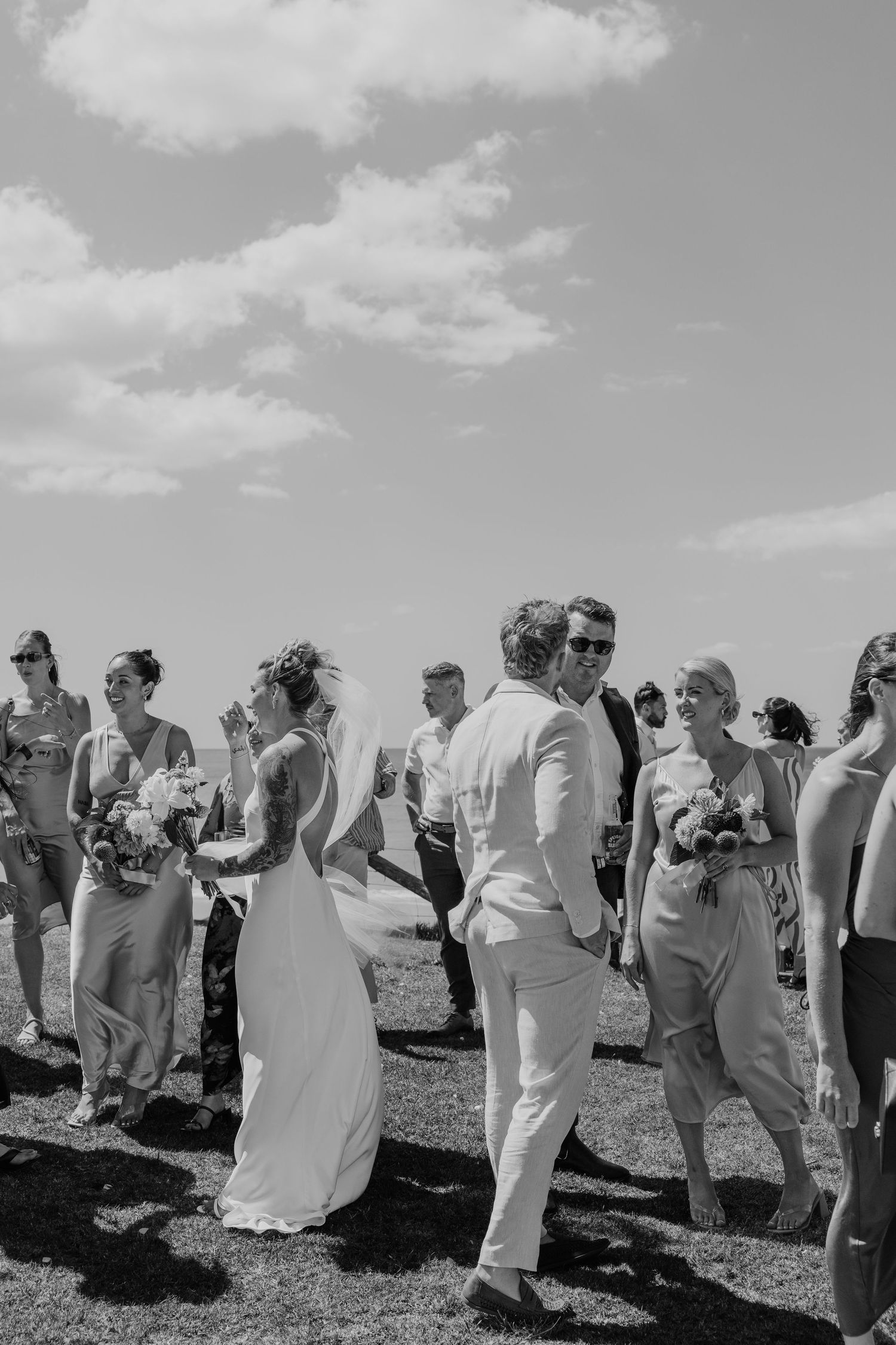 Black and white wedding photo of bride and groom walking among guests outdoors under a cloudy sky.