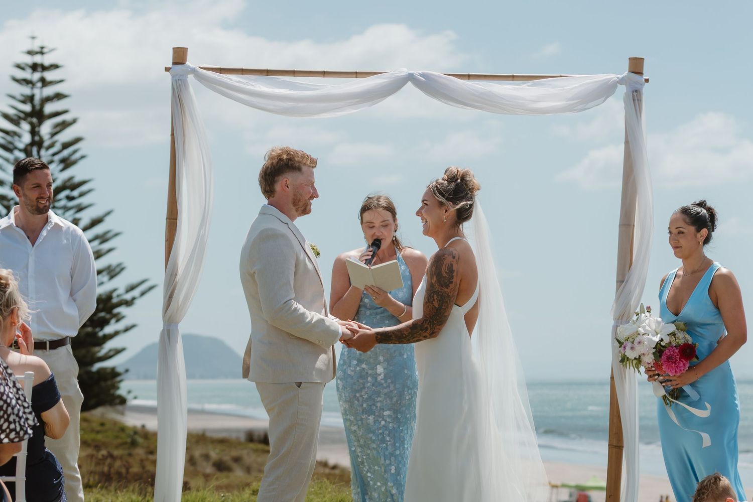 Couple exchanging vows at a romantic beach wedding ceremony under a white draped arch with ocean views.