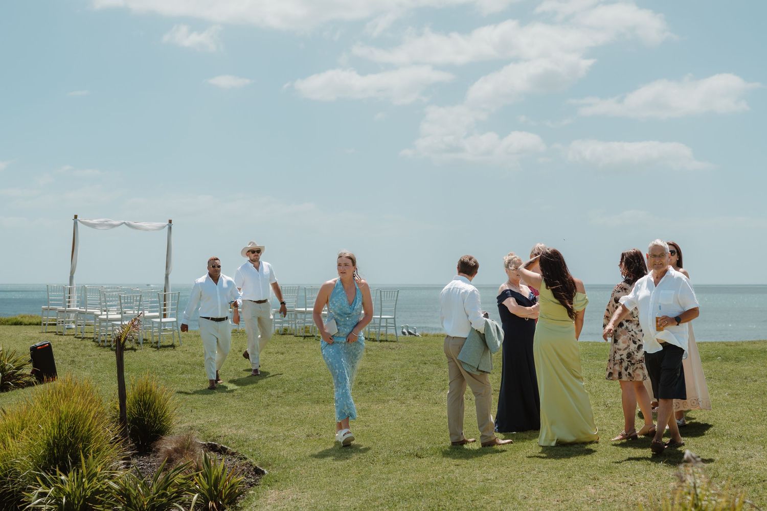Guests gather on a green lawn overlooking the ocean at an outdoor coastal wedding ceremony on a sunny day.