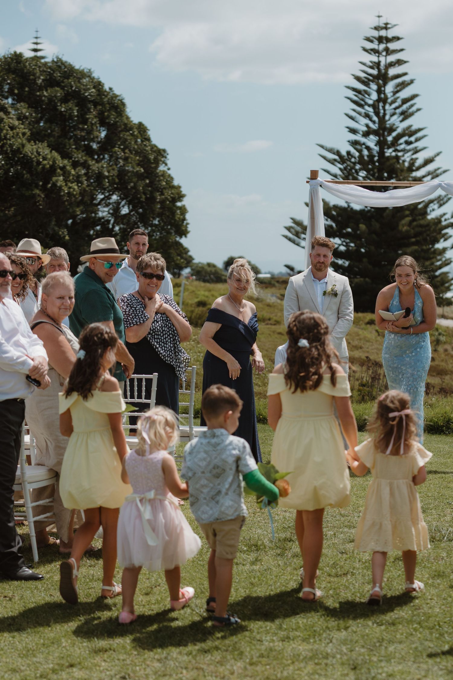 Children and guests gather outdoors at a scenic coastal wedding ceremony on a sunny day.