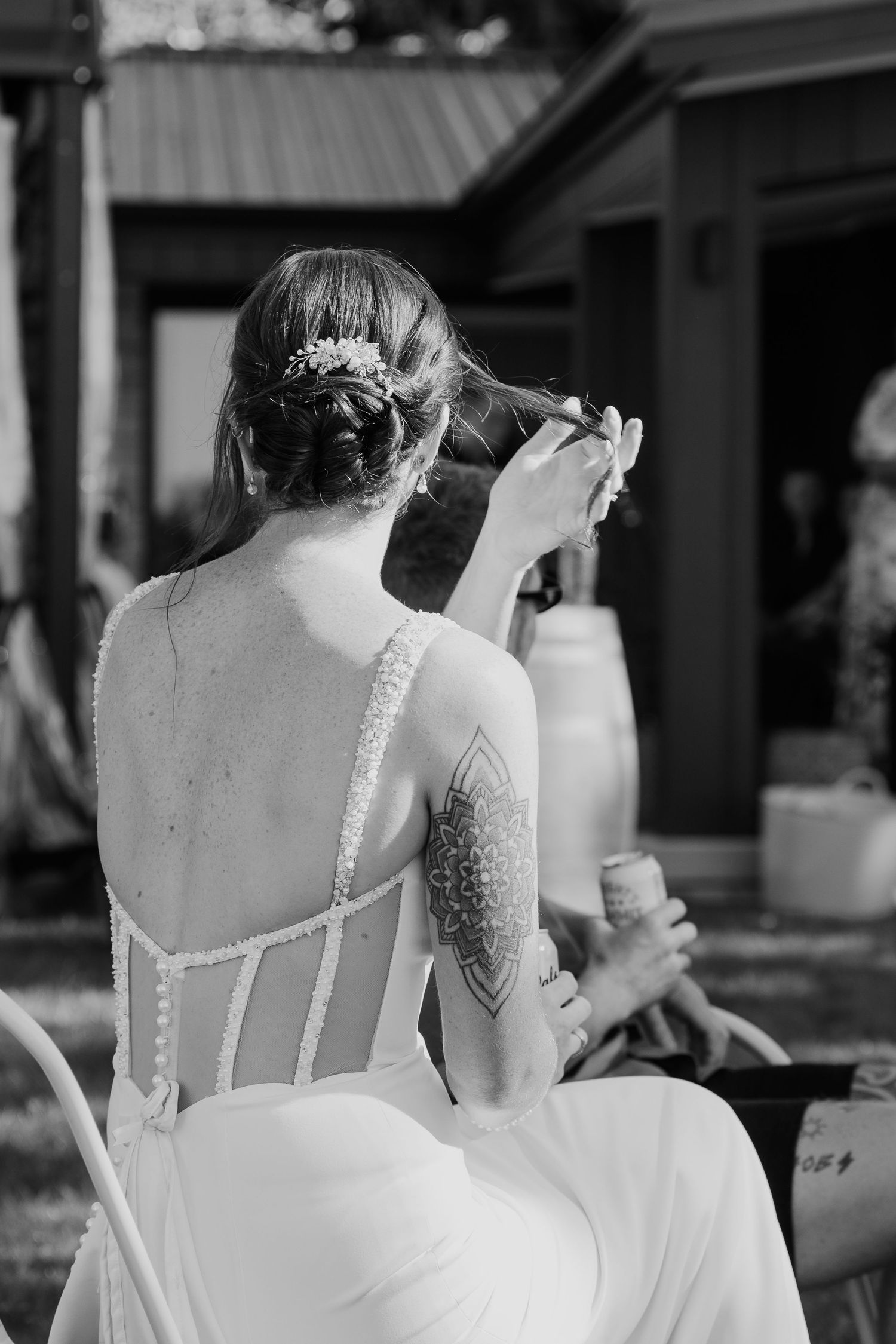 Black and white photo of a bride in a backless gown with tattoo, styling her updo hair outdoors.