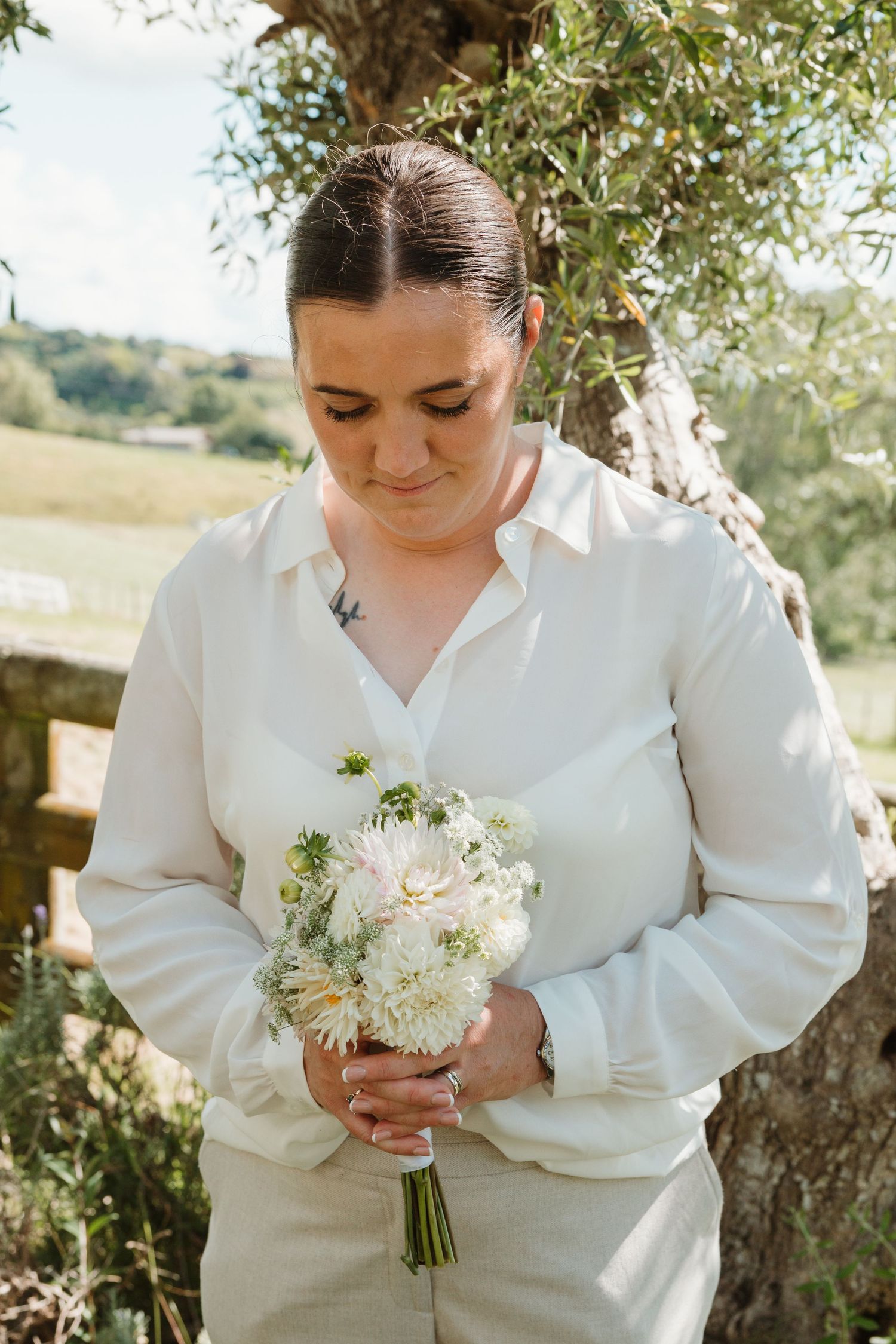 Bride in white blouse holding a small bouquet of white flowers outdoors near a tree on a sunny day.