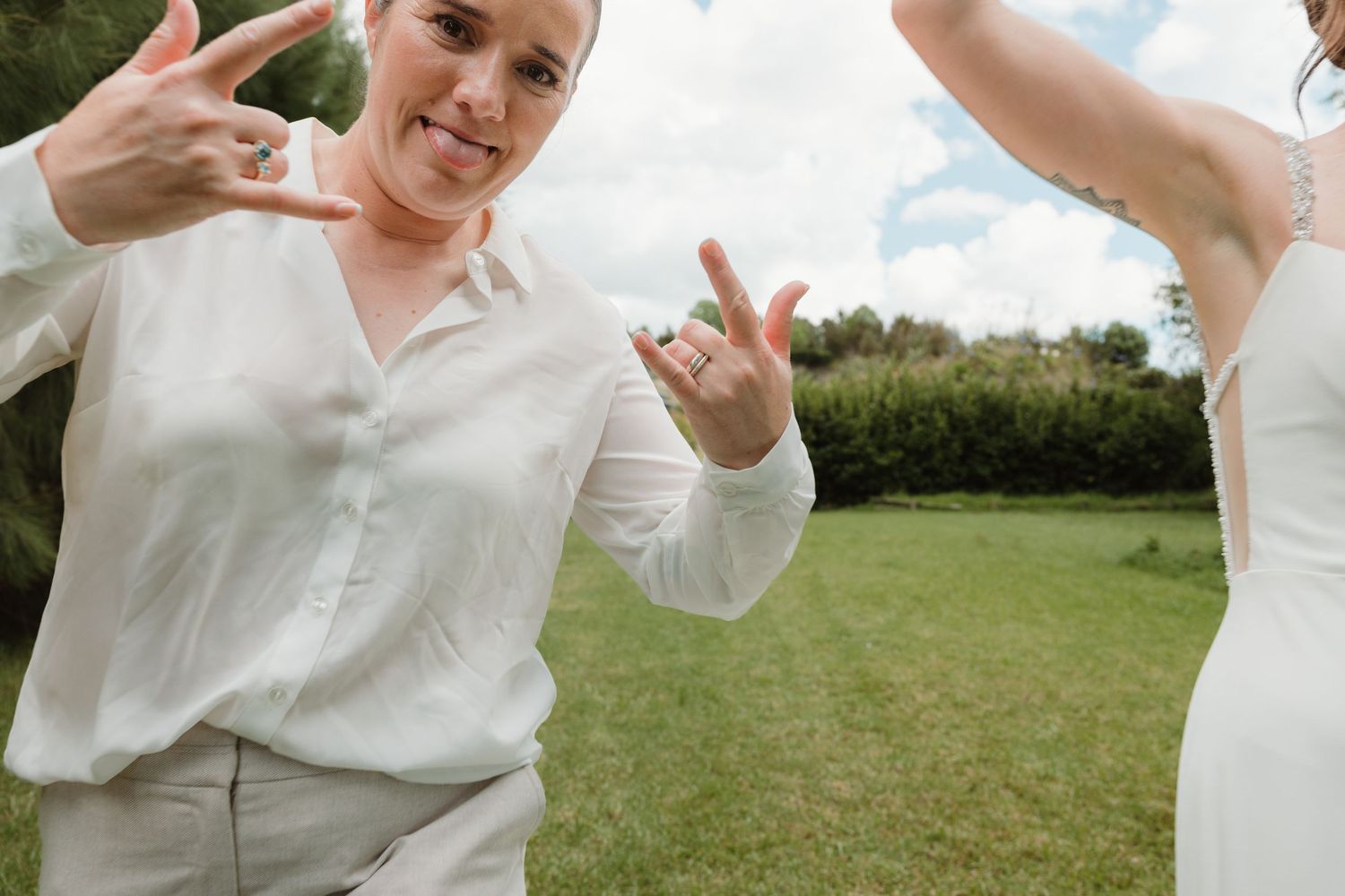 Two women showing off wedding rings outdoors, smiling and making hand gestures on a green lawn.