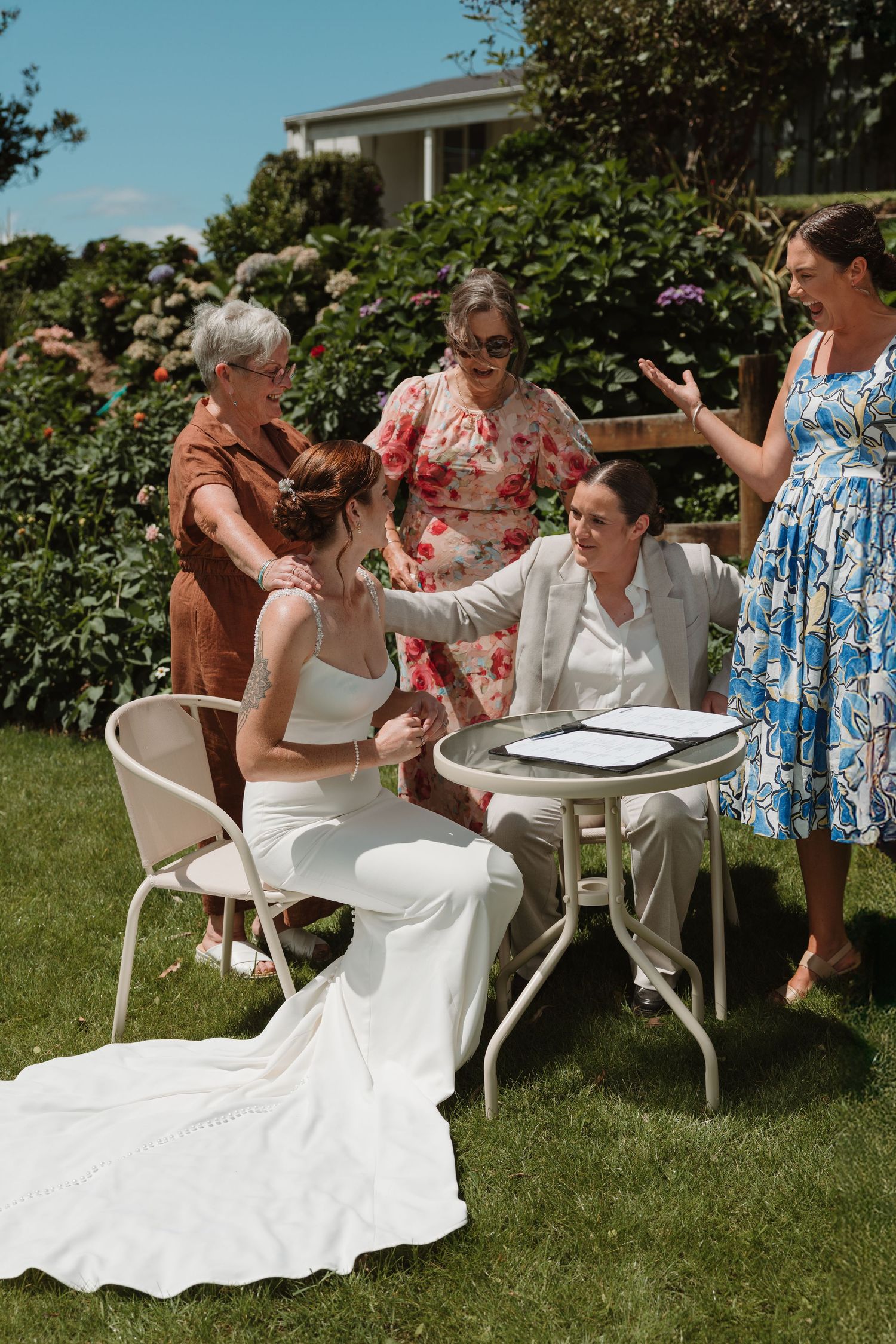 Two brides sign wedding documents outdoors surrounded by smiling guests in a sunny garden setting.