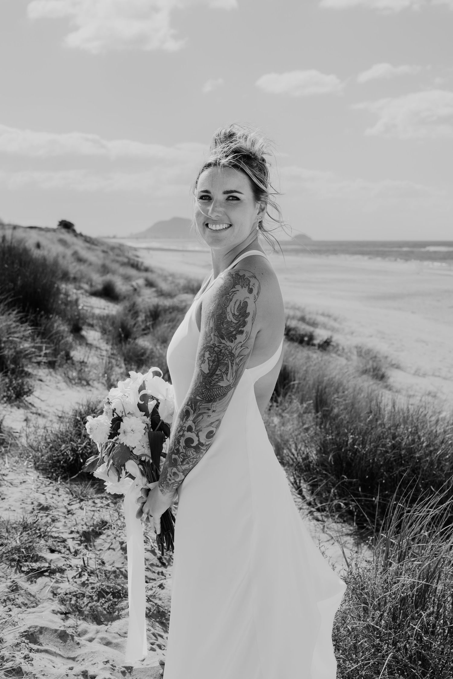 Black and white photo of a tattooed bride in a white gown holding a bouquet on a sandy beach.
