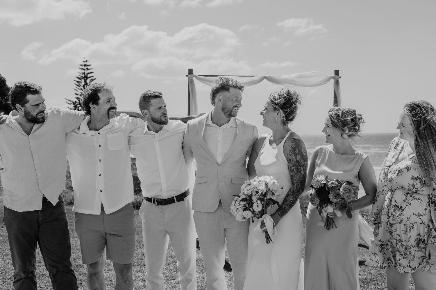 Black and white photo of a wedding party laughing together outdoors near a rustic arch on a scenic coastal backdrop.
