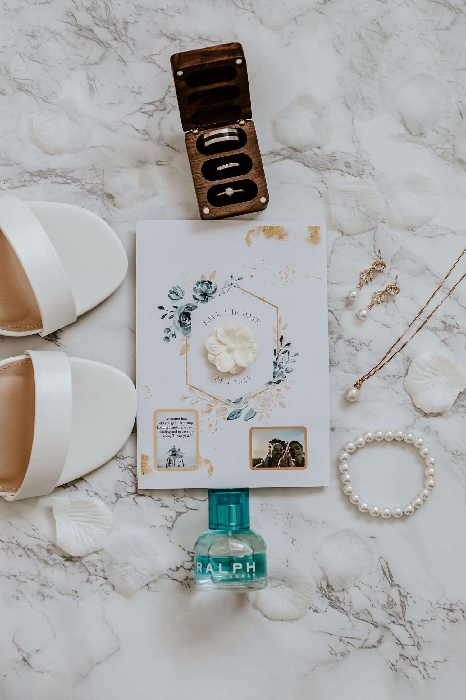 Wedding accessories including white shoes, invitation with floral design, turquoise nail polish and pearl bracelet on marble surface.