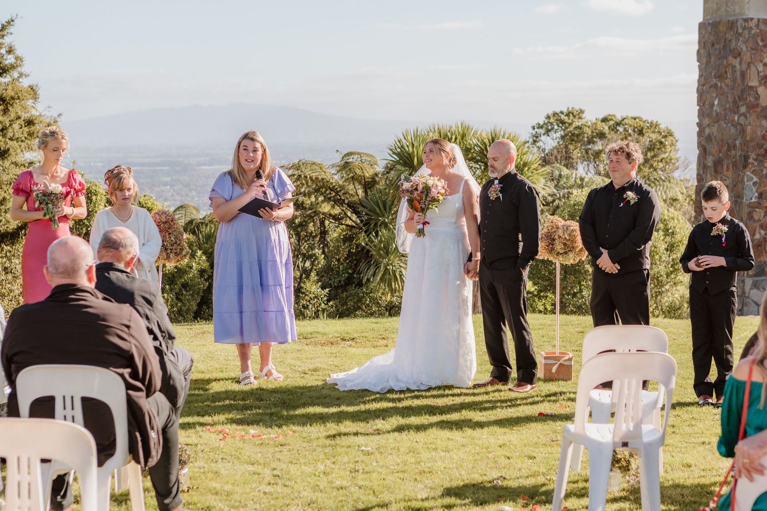 An outdoor wedding ceremony takes place on a scenic hilltop with white chairs and beautiful mountain views in the background.