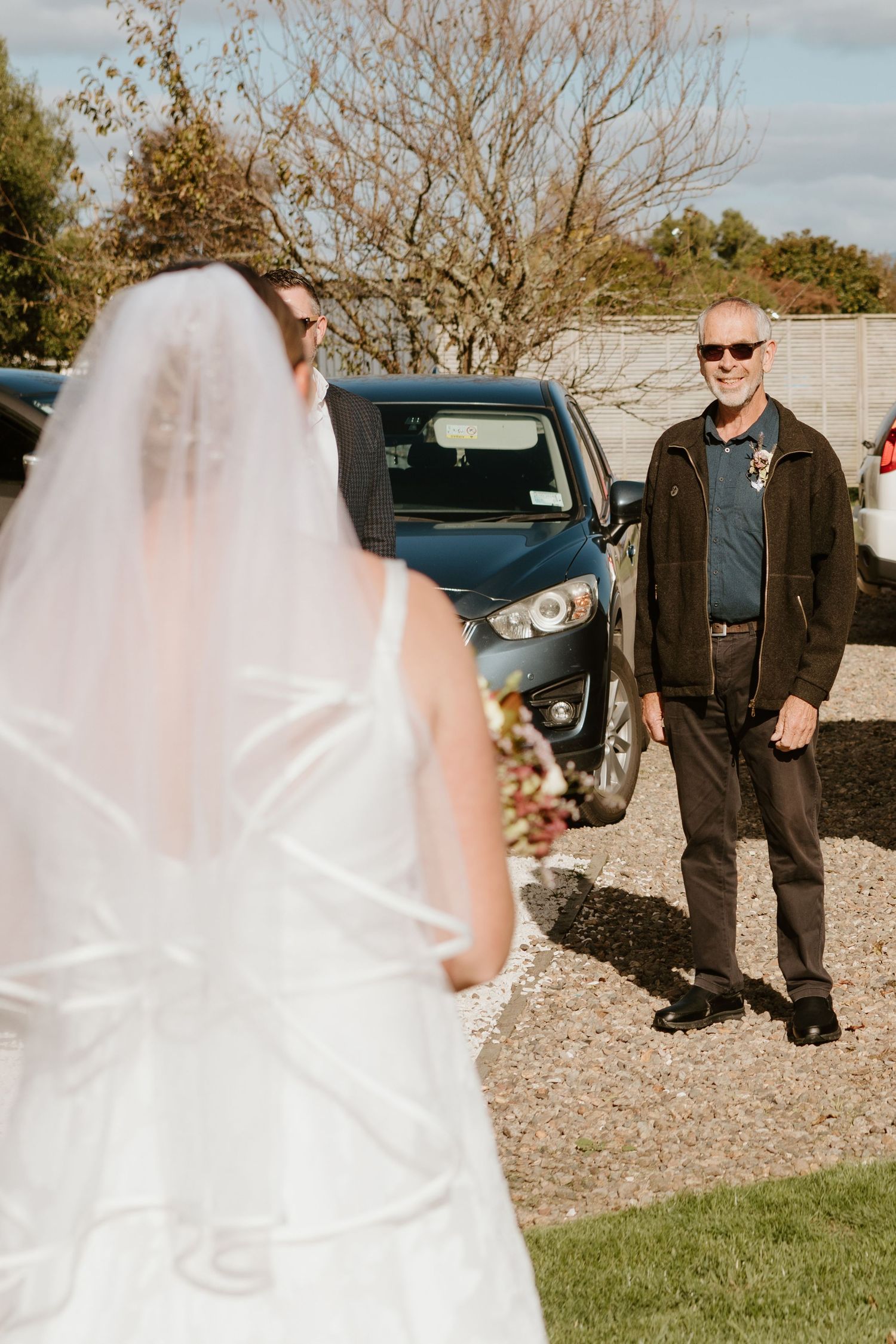 A bridal veil flows in the breeze on a sunny day while standing on grassy terrain near parked cars.