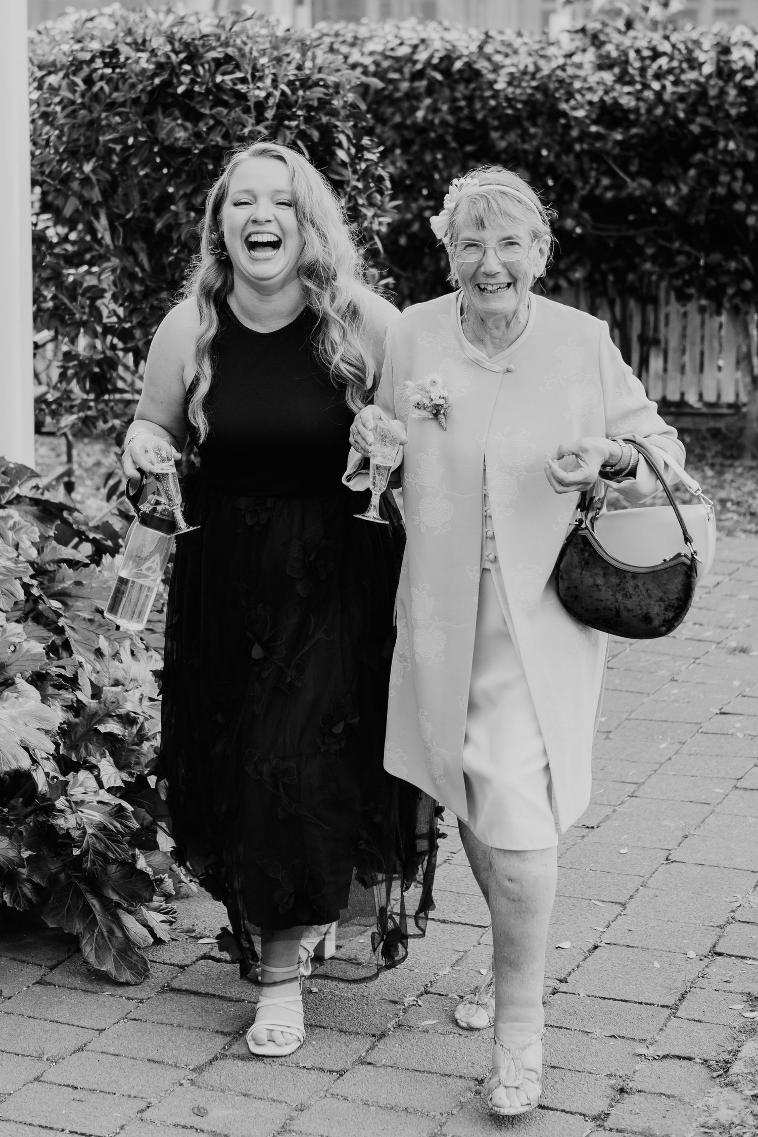 Black and white photograph showing two people walking and laughing together on a garden path with hedges in the background.