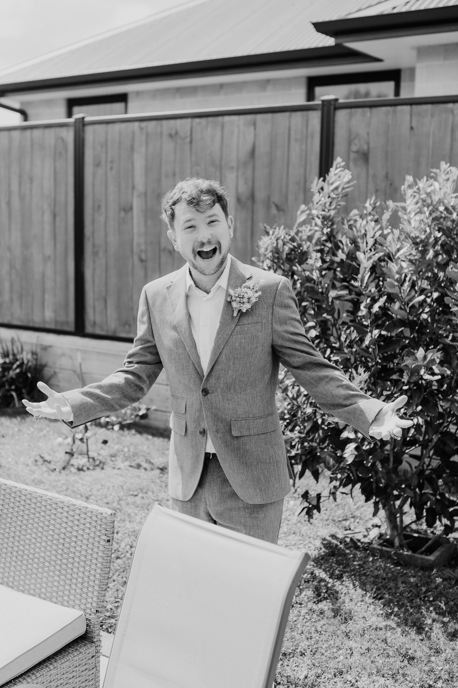 A person in a gray suit and boutonniere laughs joyfully in a backyard garden setting with a wooden fence background.