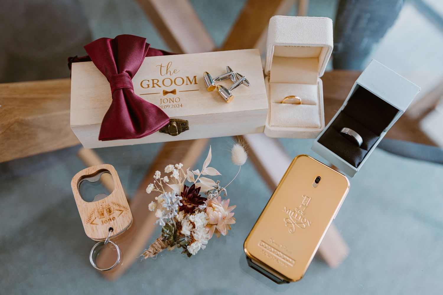 Wedding accessories including a burgundy bow tie, ring boxes, floral arrangement, bottle opener and gold phone displayed on blue fabric.