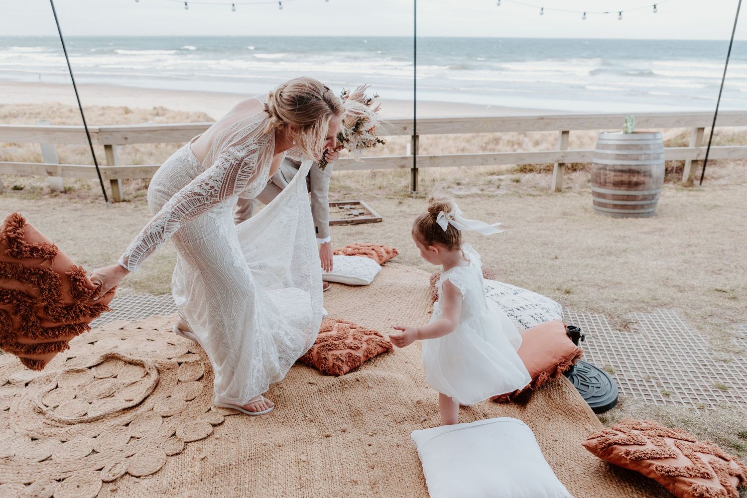 Two people in white dresses share a sweet moment while sitting on blankets at a windy beach.