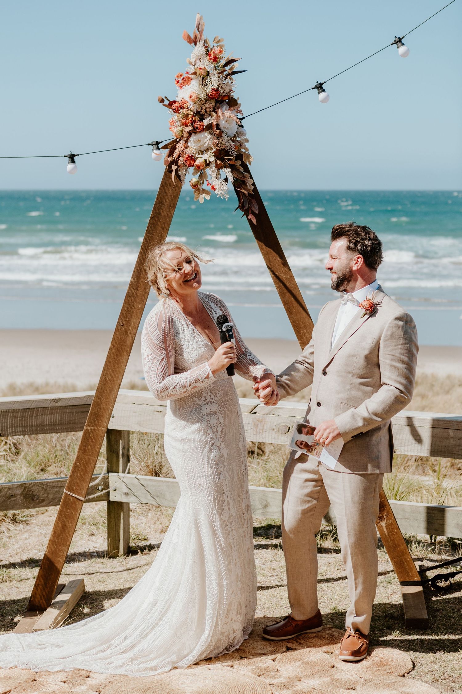 Rustic beach wedding ceremony setup with wooden arch adorned with dried flowers against turquoise ocean backdrop.