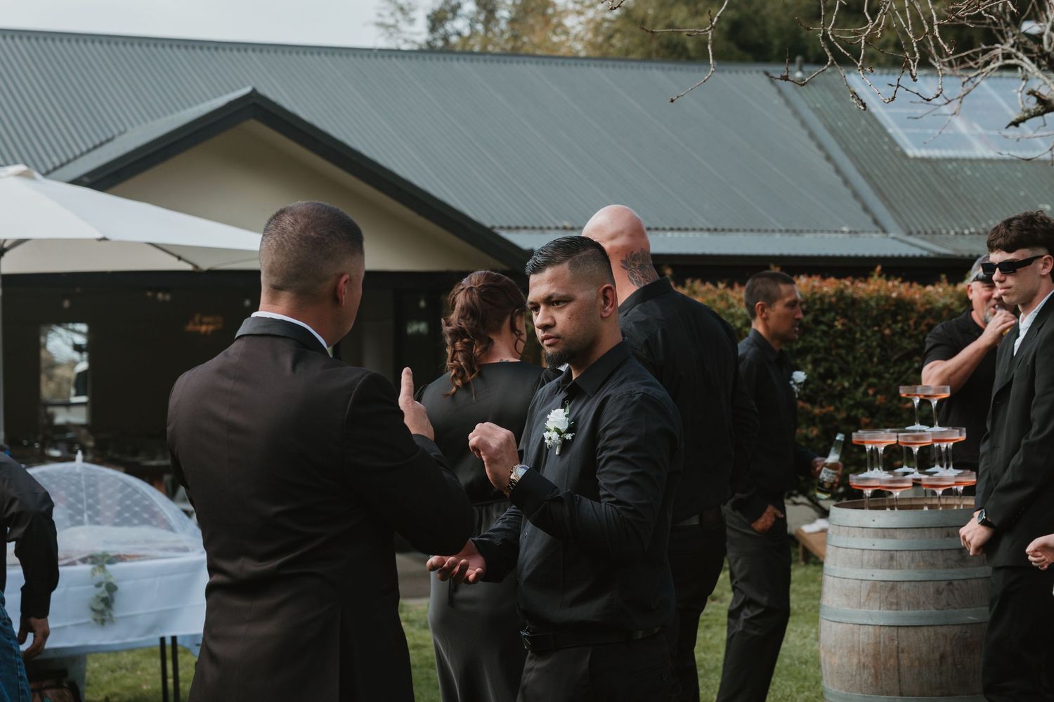 Wedding guests clink glasses in a celebratory toast at an outdoor reception venue with rustic wine barrels.