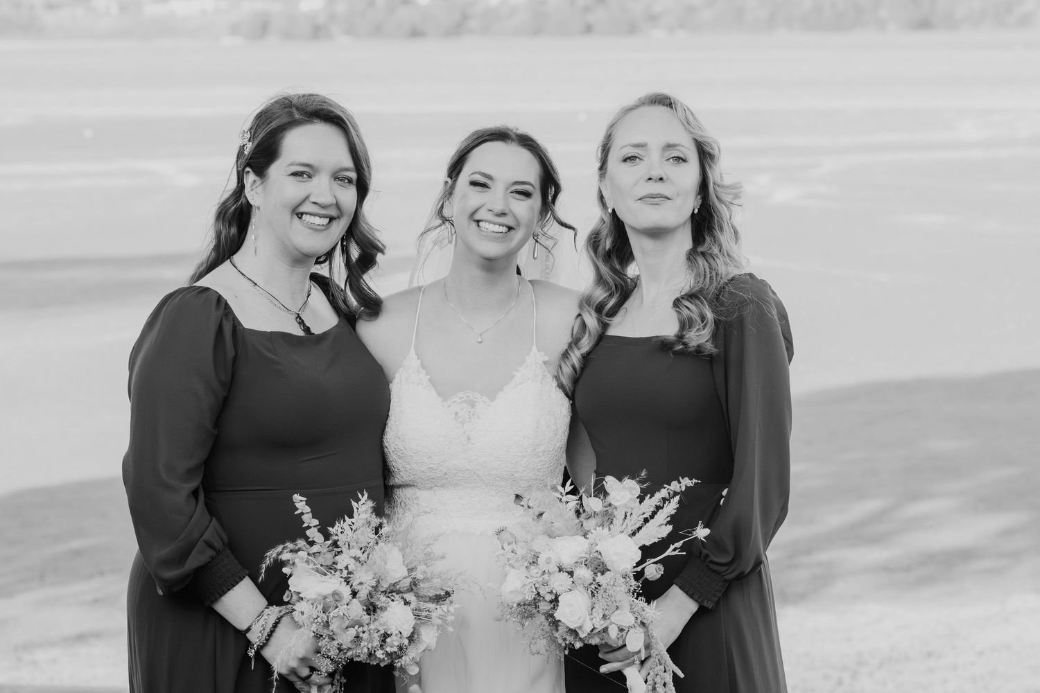 A bride in white poses with two bridesmaids in dark dresses holding floral bouquets.