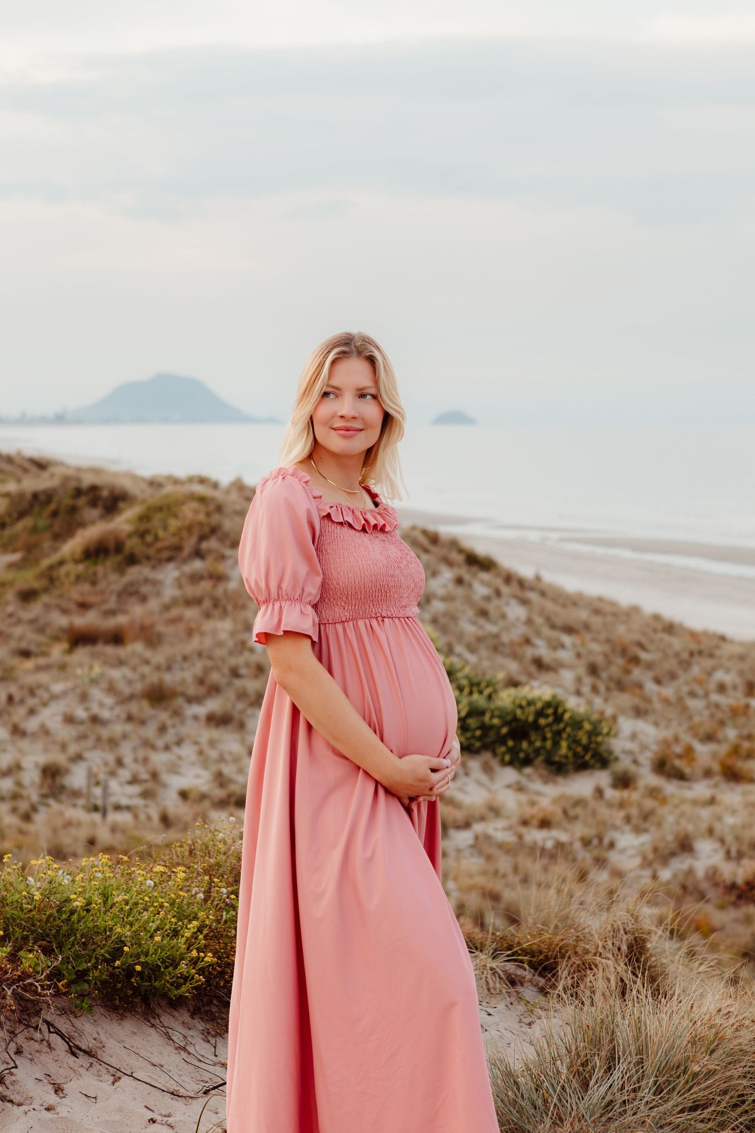 Woman in pink maxi dress stands on coastal dunes with ocean and mountains in background at sunset. Maternity session Tauranga, Papamoa