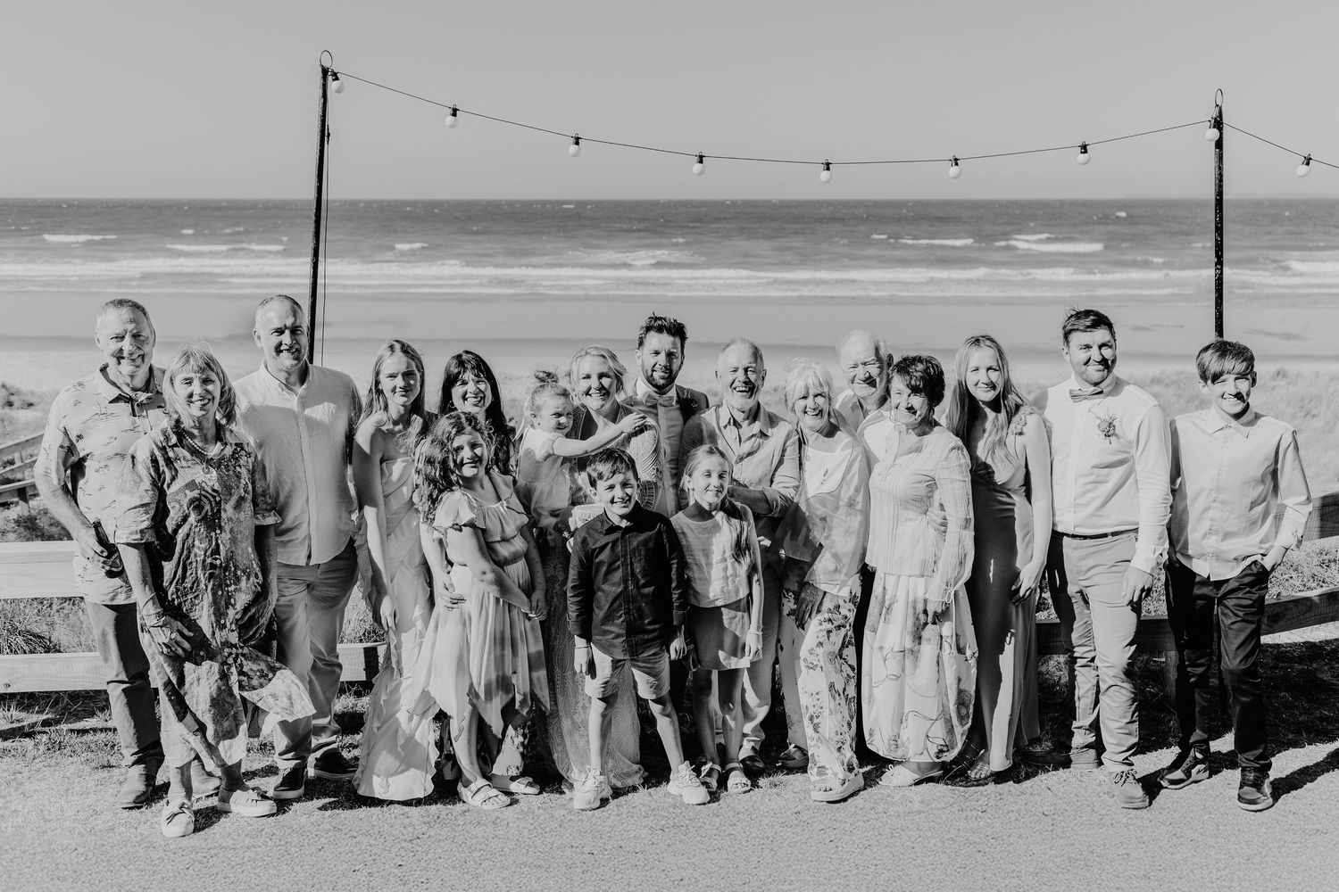 A large wedding group stands together on a beach under string lights with the ocean visible behind them.