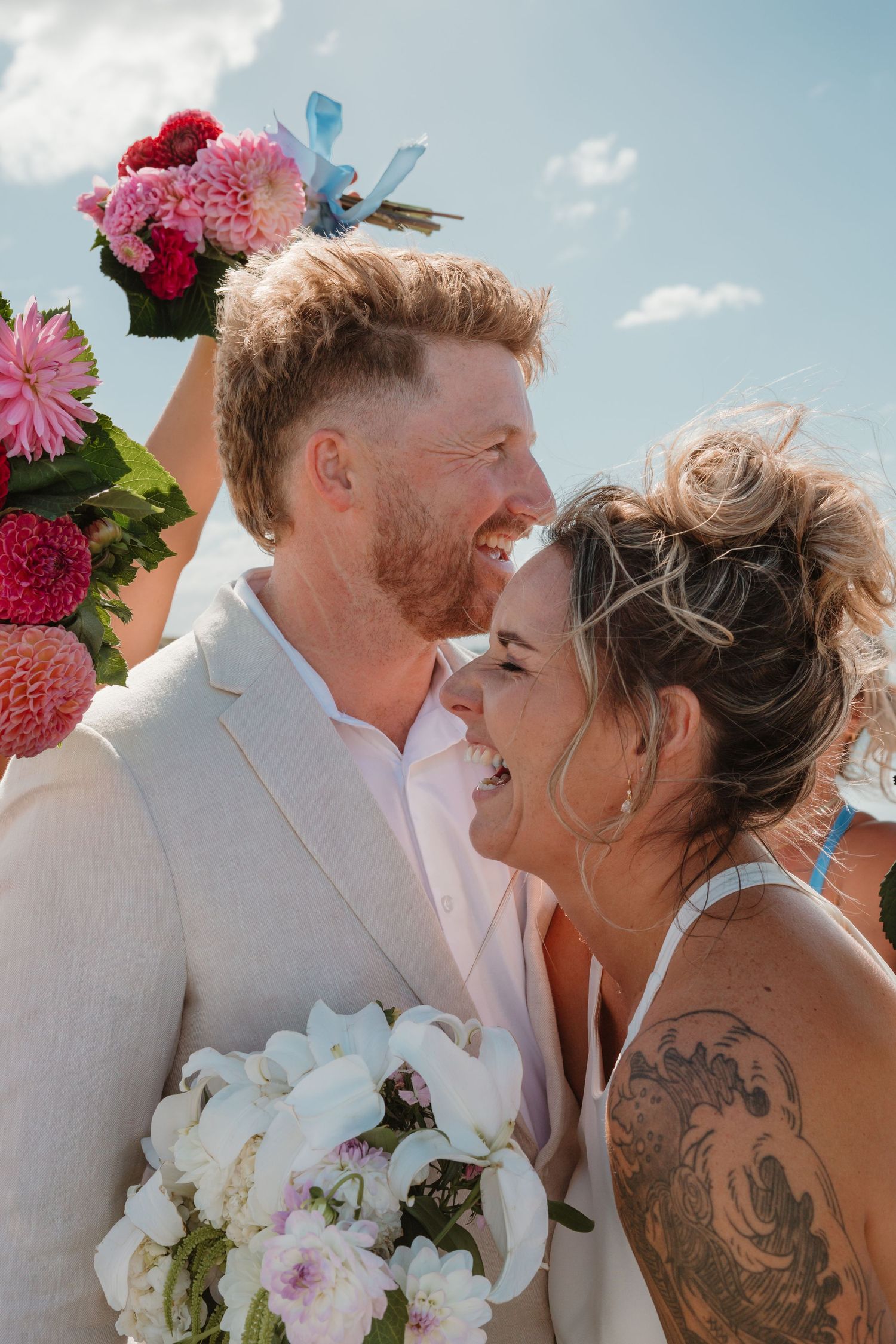 A joyful couple shares a candid moment at their beach wedding with pink and white floral bouquets against a blue sky.