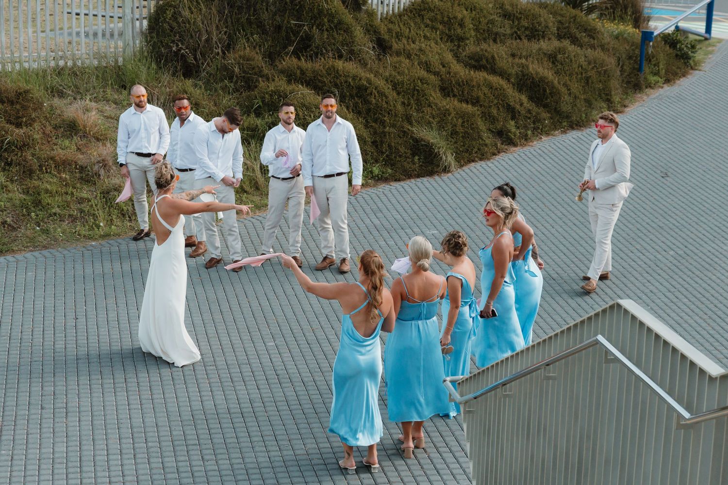 Wedding party in blue bridesmaid dresses and white suits pose on a coastal pathway during an outdoor beach wedding celebration.